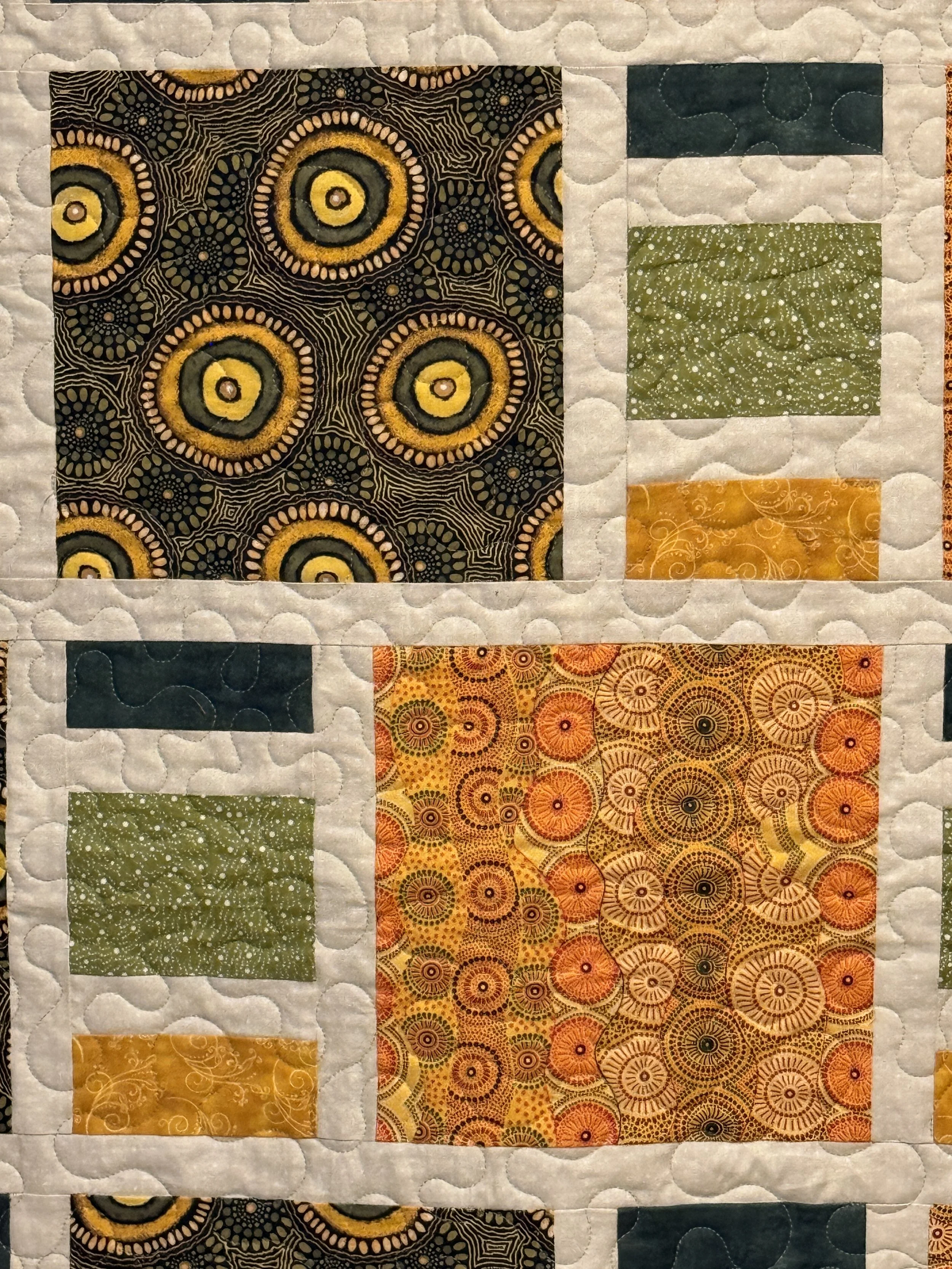 Close-up of a quilt featuring multiple fabric patterns, including circular, spiral, and geometric designs in colors such as yellow, orange, green, and dark blue. The quilt has a white background with quilting stitching visible.