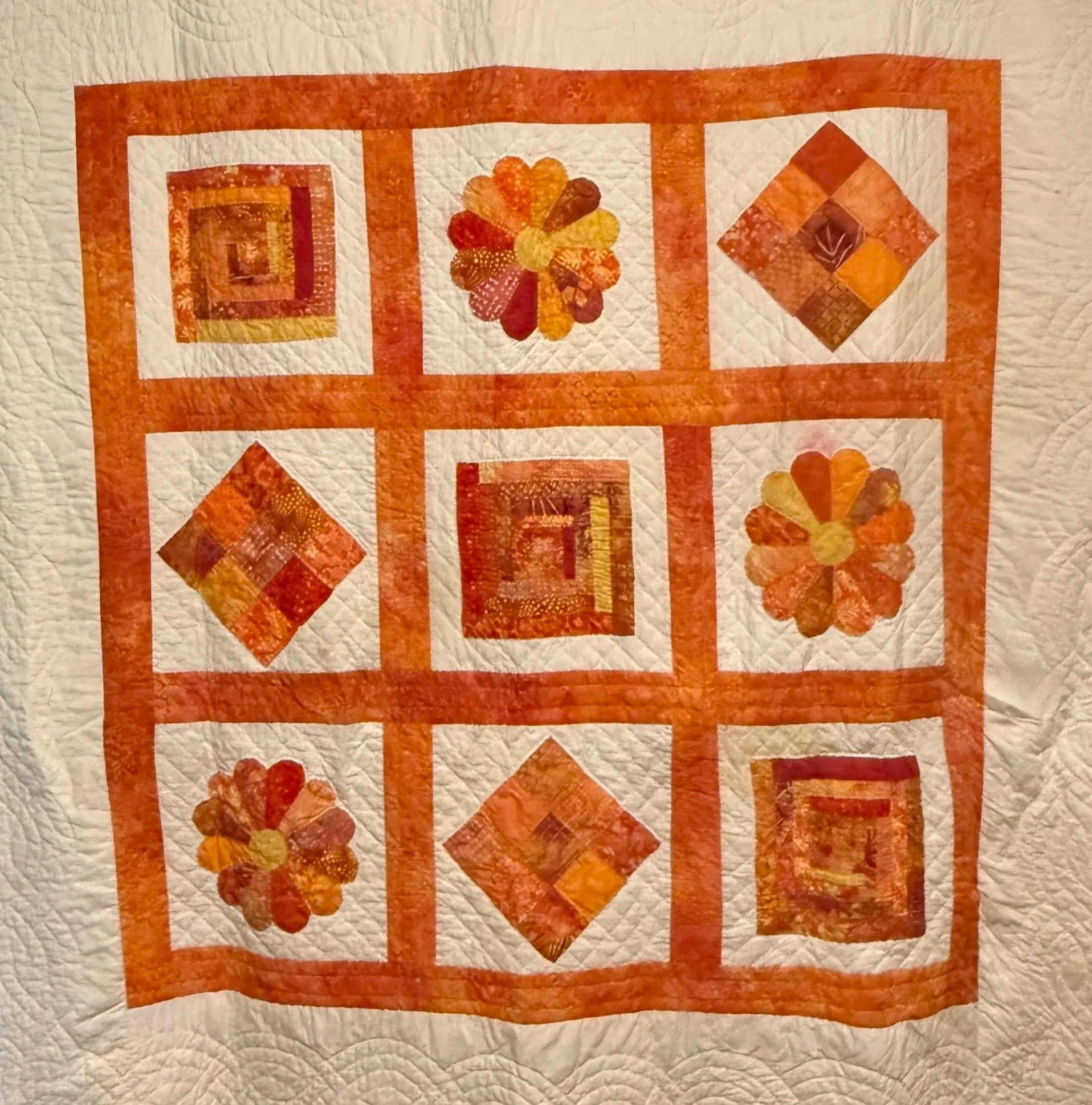 A quilt with a pattern of nine blocks arranged in a 3x3 grid, featuring floral, square, and diamond shapes in warm orange, yellow, and red tones.