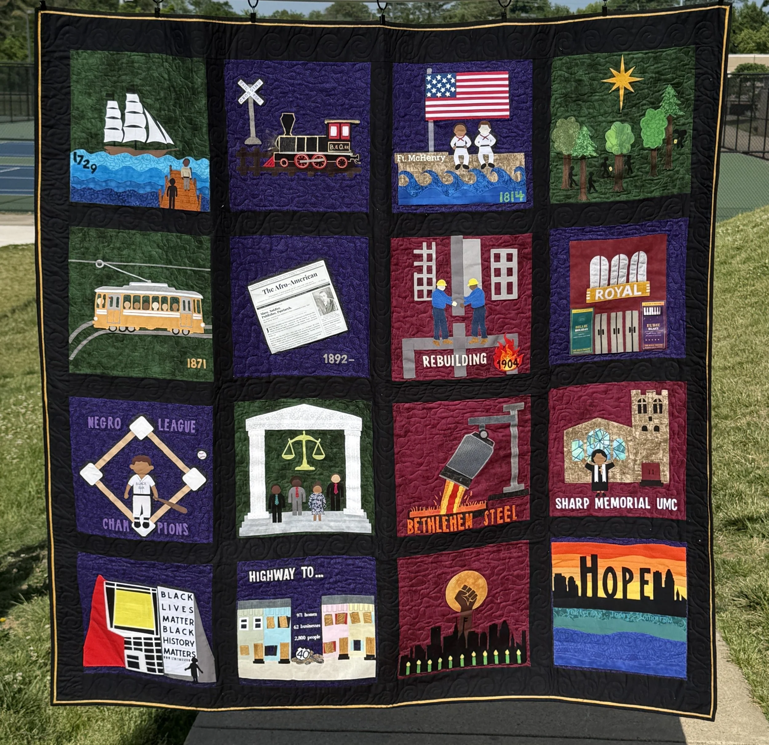 A quilt with various patches depicting historical and cultural themes, including a sailing ship, a train, an American flag with two children saluting, trees, a tram, the Black Lives Matter sign, and cityscape buildings, all bordered with black fabric