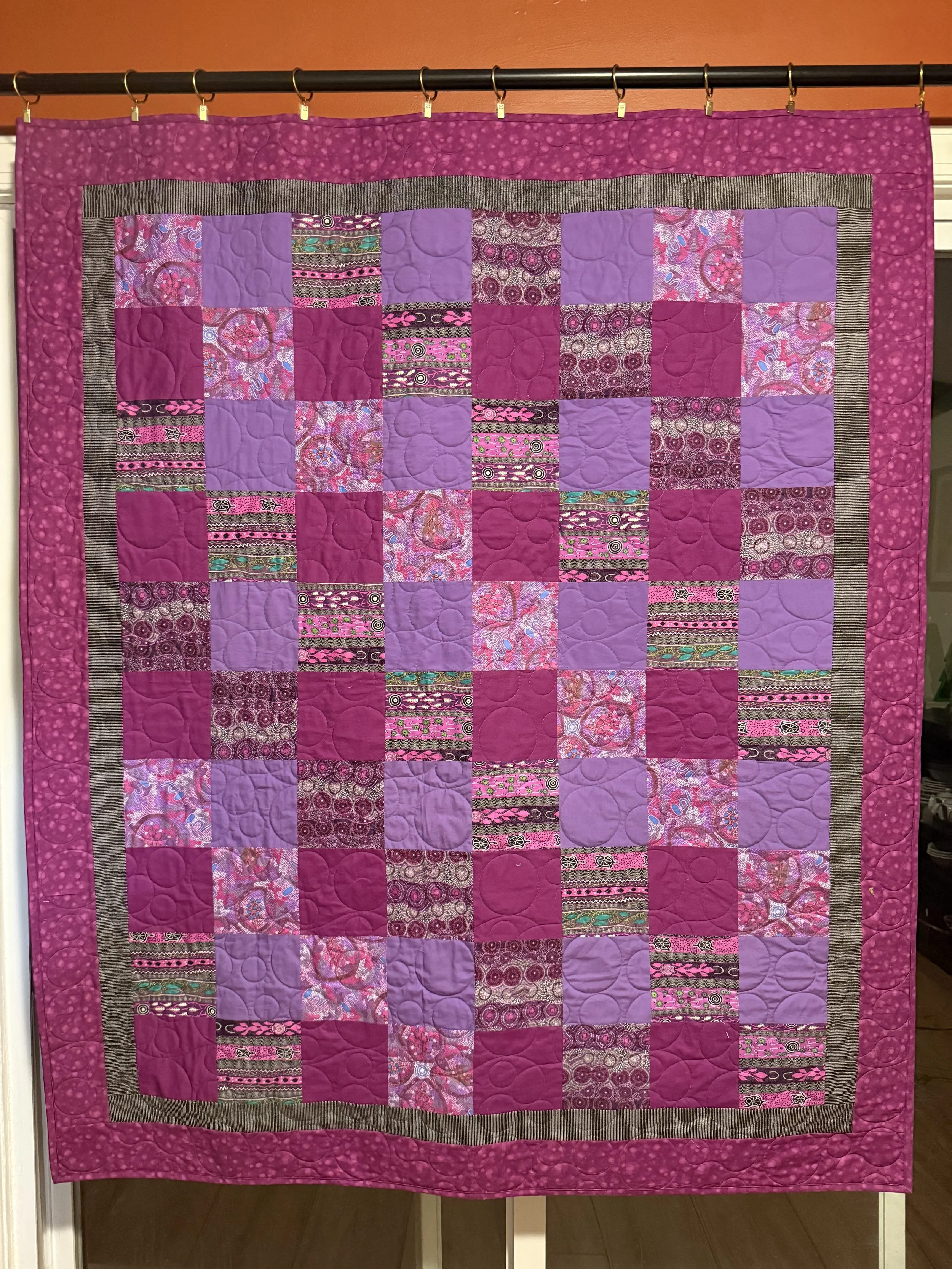 A colorful patchwork quilt with various pink, purple, and patterned fabric squares, bordered by a pink patterned outer border and hanging on a black rod.