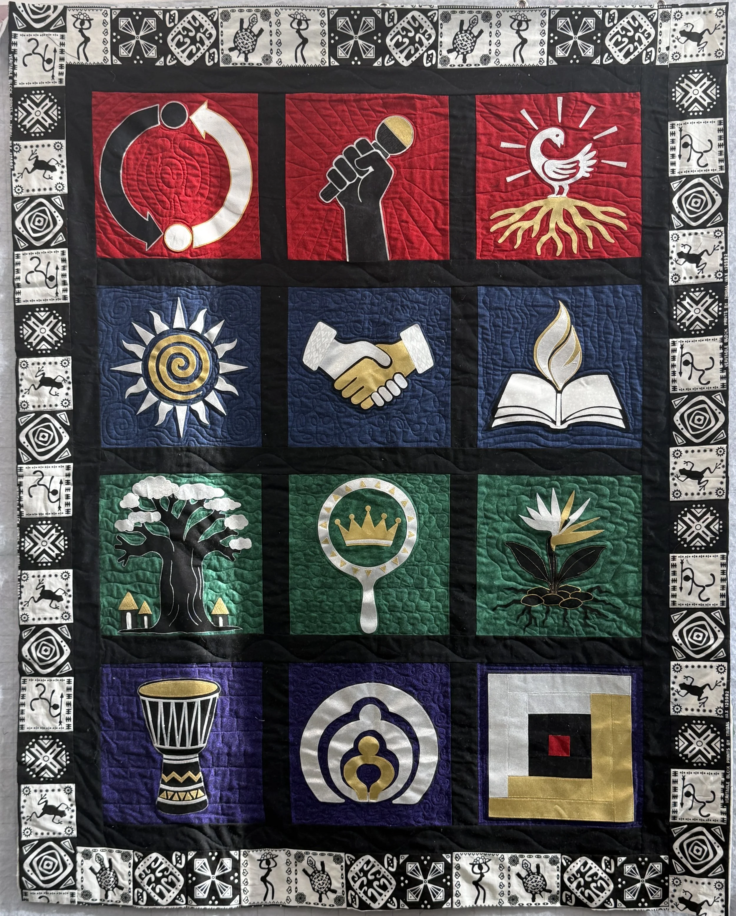 A fabric quilt with various symbols and icons depicting social and political themes. The top row contains black and white patterns with stylized animals and geometric designs. The main sections display symbols such as a compact disc with arrows, a clenched fist with a microphone, a rooster on roots, a sun with a spiral center, a handshake, an open book with a flame, a drum, a crown, and a flower, all in bold colors including red, blue, green, white, black, and gold. The quilt is bordered with black and white patterned tabs featuring stylized animals and geometric motifs.