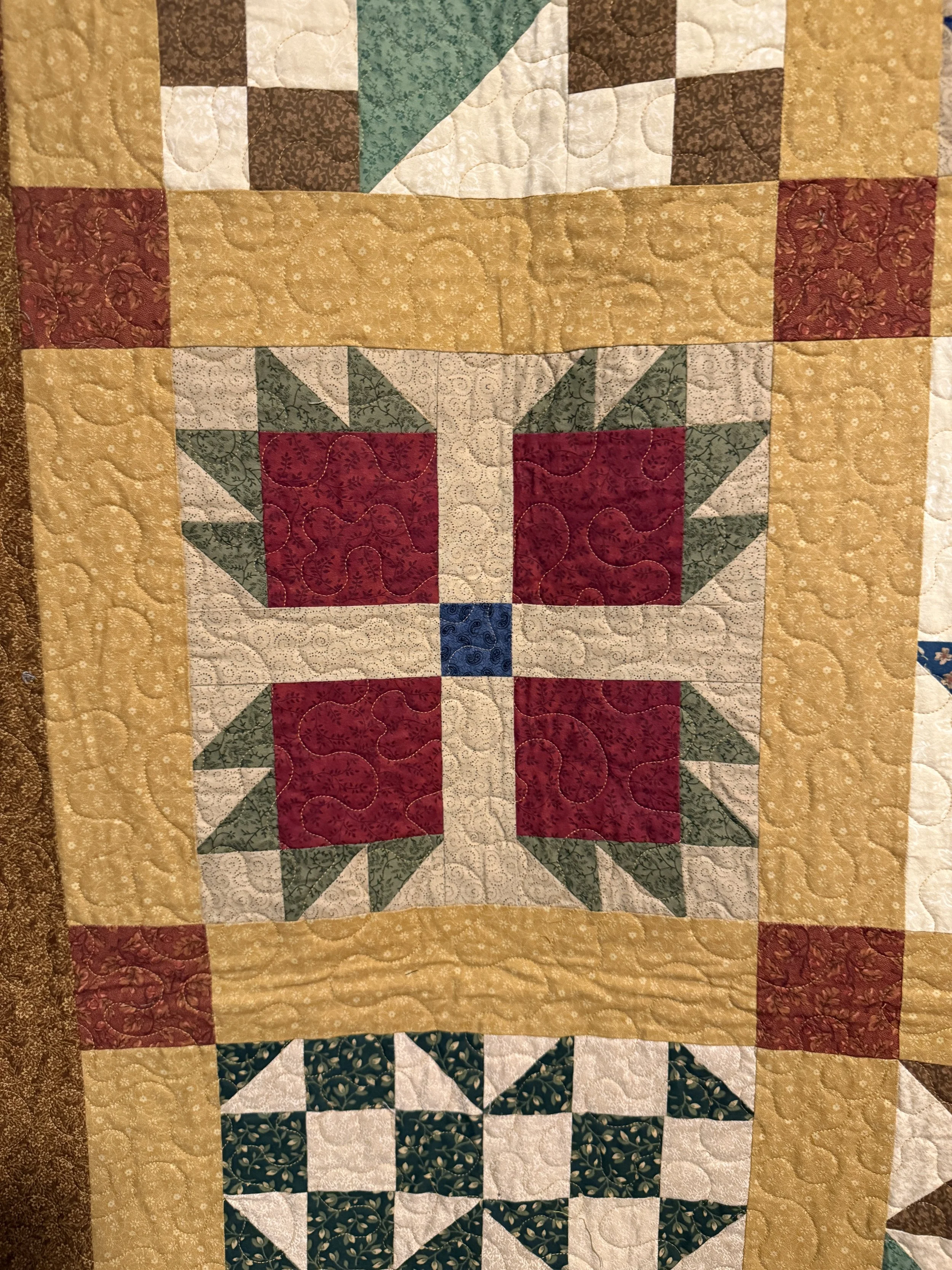 Close-up of a quilt with a central square featuring red, white, green, and blue fabric, surrounded by yellow, brown, green, and black patterned borders and designs.