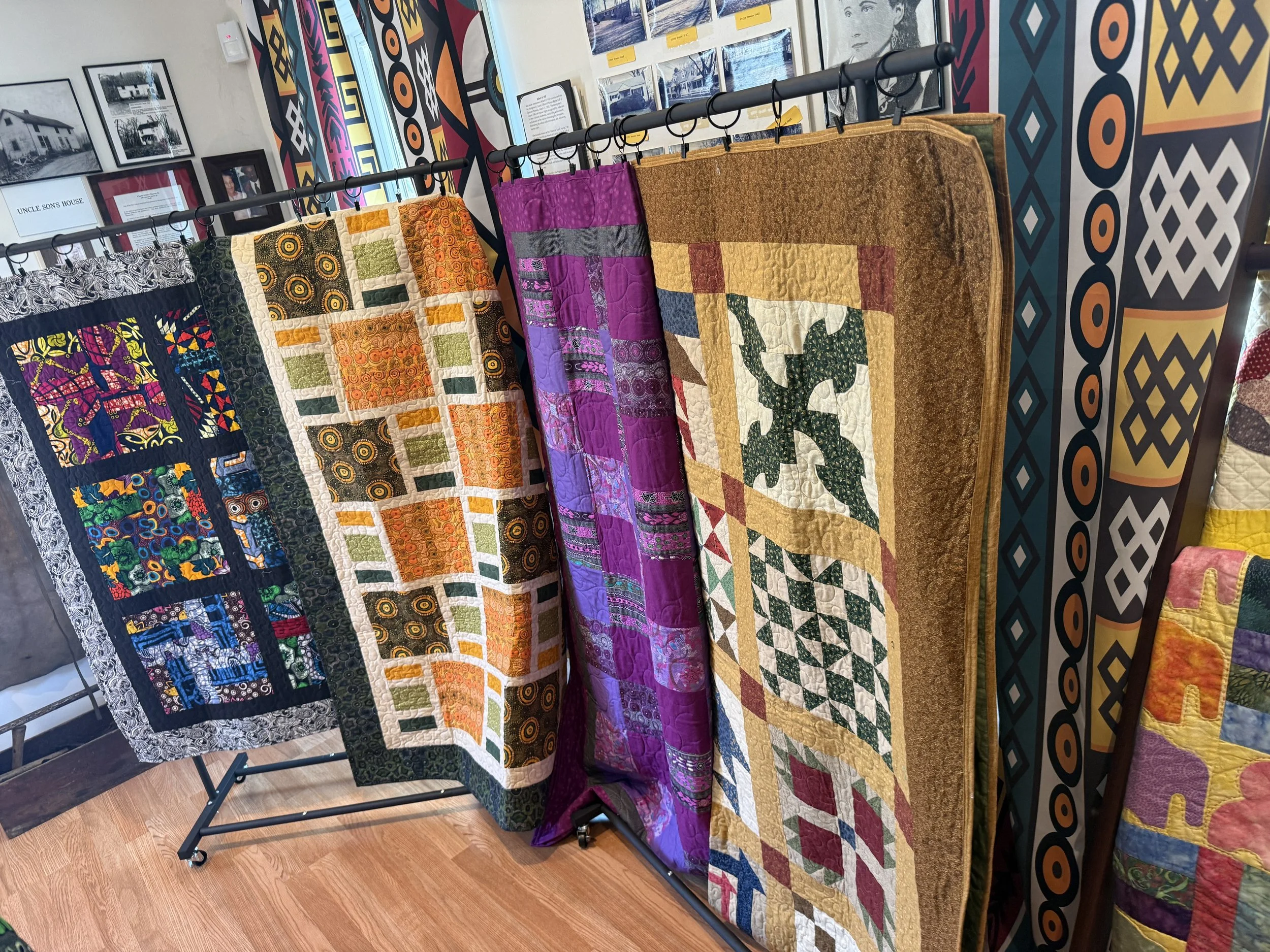 Display of colorful quilts hanging on a rack in a room with decorated walls, framed pictures, and colorful patterns.