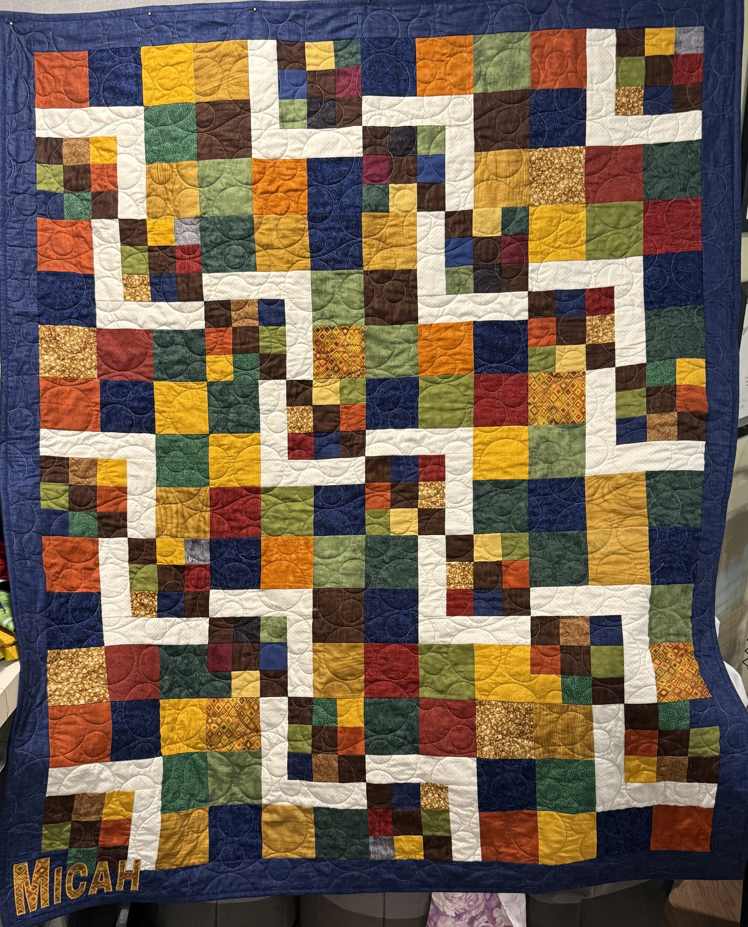 A colorful quilt made of small fabric squares arranged in a pattern with elements of red, yellow, green, white, blue, and brown, with a blue border and the name 'MICHIGAN' embroidered in the lower left corner.