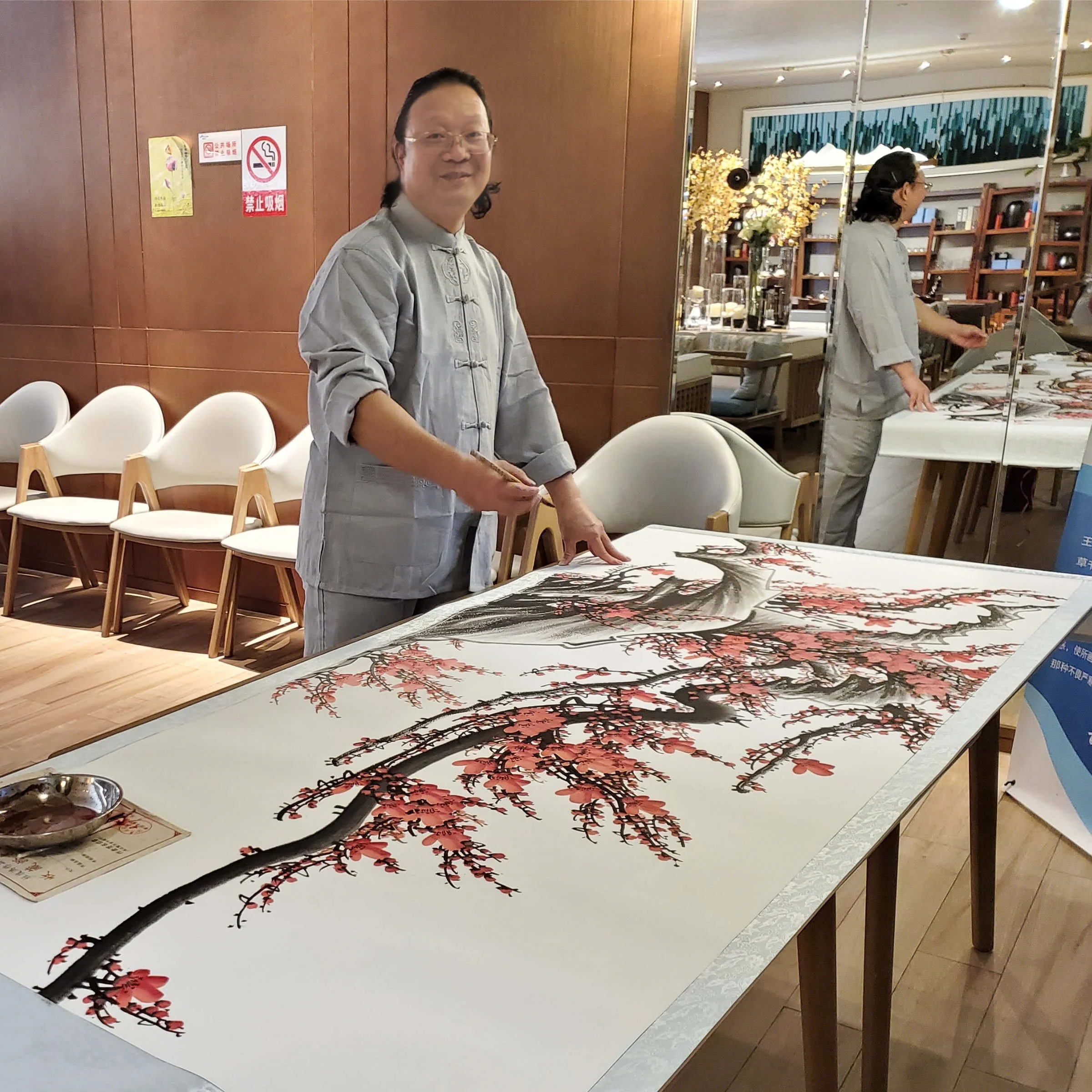Artist standing next to a large traditional Chinese ink painting of a cherry blossom tree on a table, with a mirror in the background reflecting her and the painting.