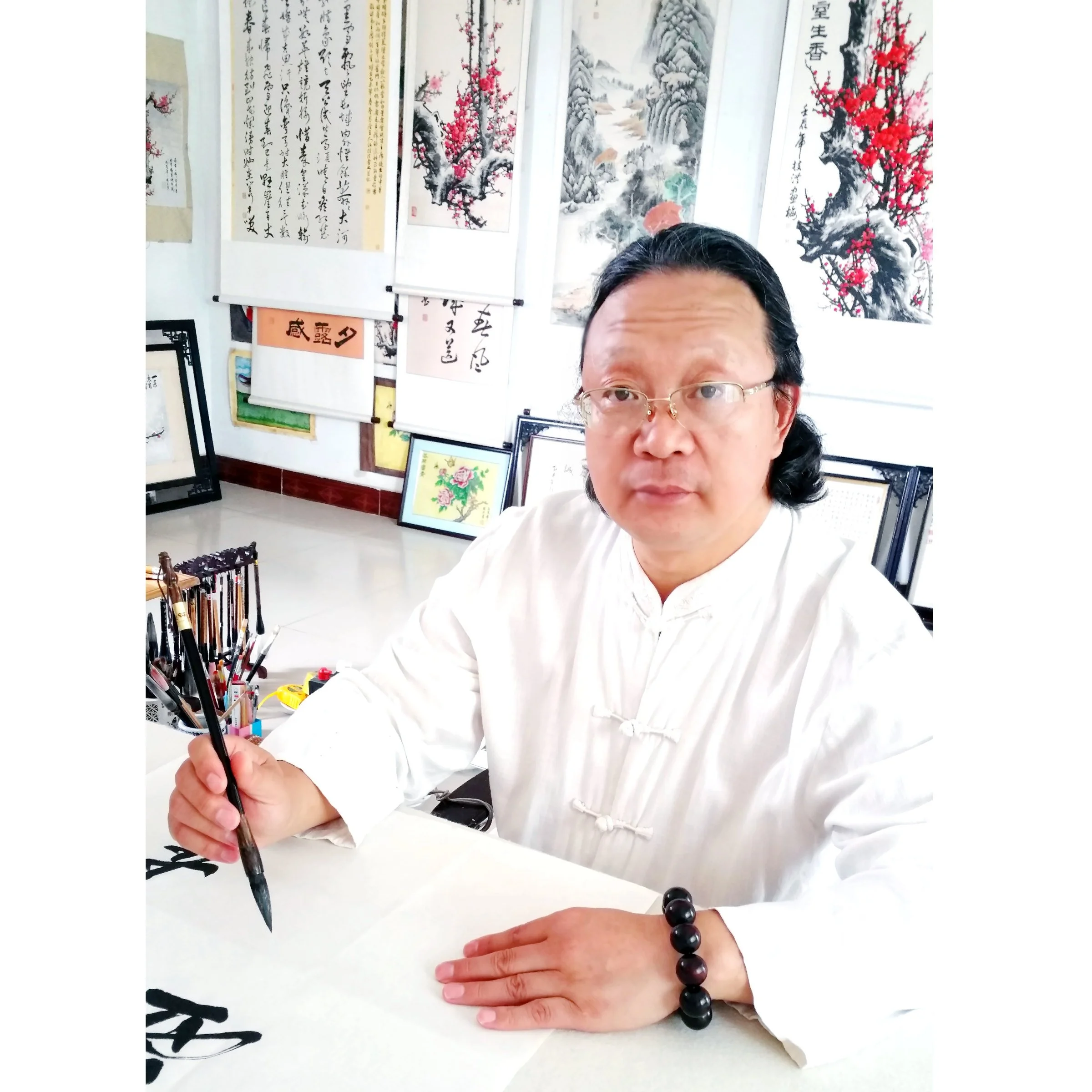 Artist practicing Chinese calligraphy at a desk, holding a calligraphy brush, with traditional Chinese paintings and calligraphy scrolls displayed on the wall in the background.