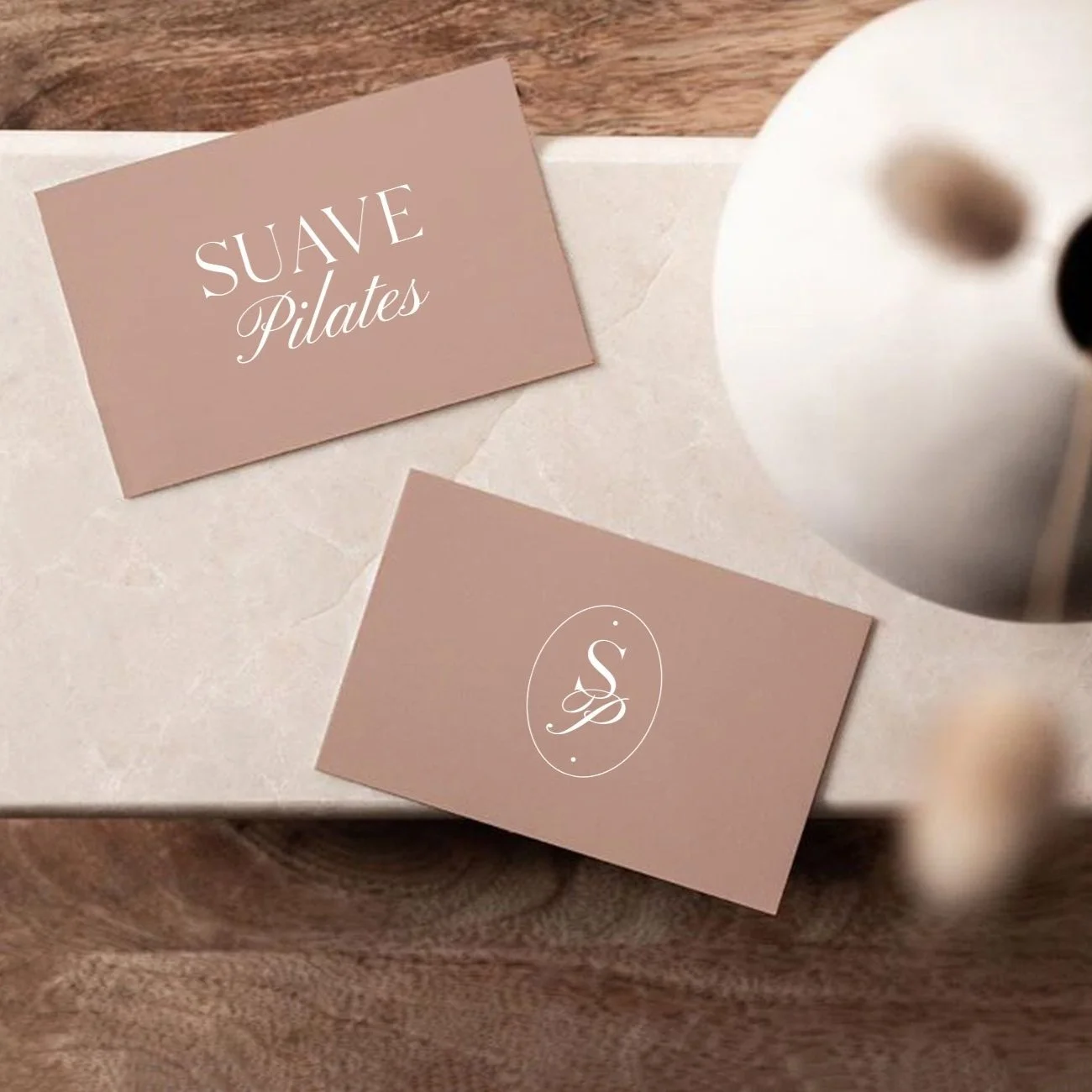 Business cards on a white marble tray, with one reading 'Suave Pilates' and the other featuring a stylized 'S' logo, next to a white object, possibly a vase.