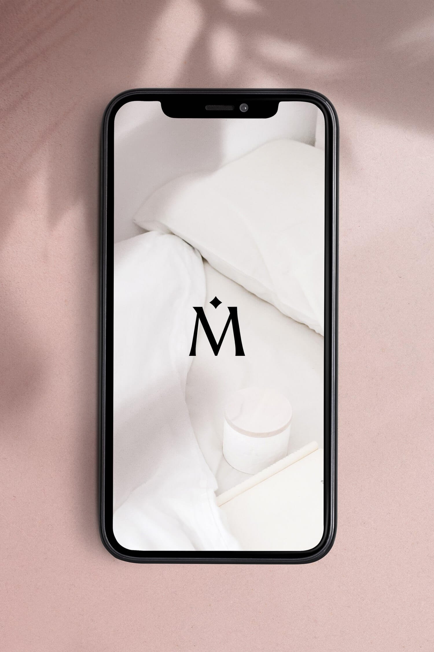 A smartphone displaying a minimalist wallpaper with a black letter M and a small diamond shape above it, set against a white bed and pillow background.