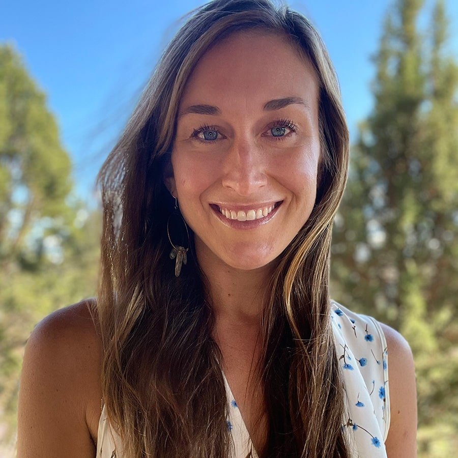 A woman with long brown hair and blue eyes smiling outdoors with trees and a blue sky in the background