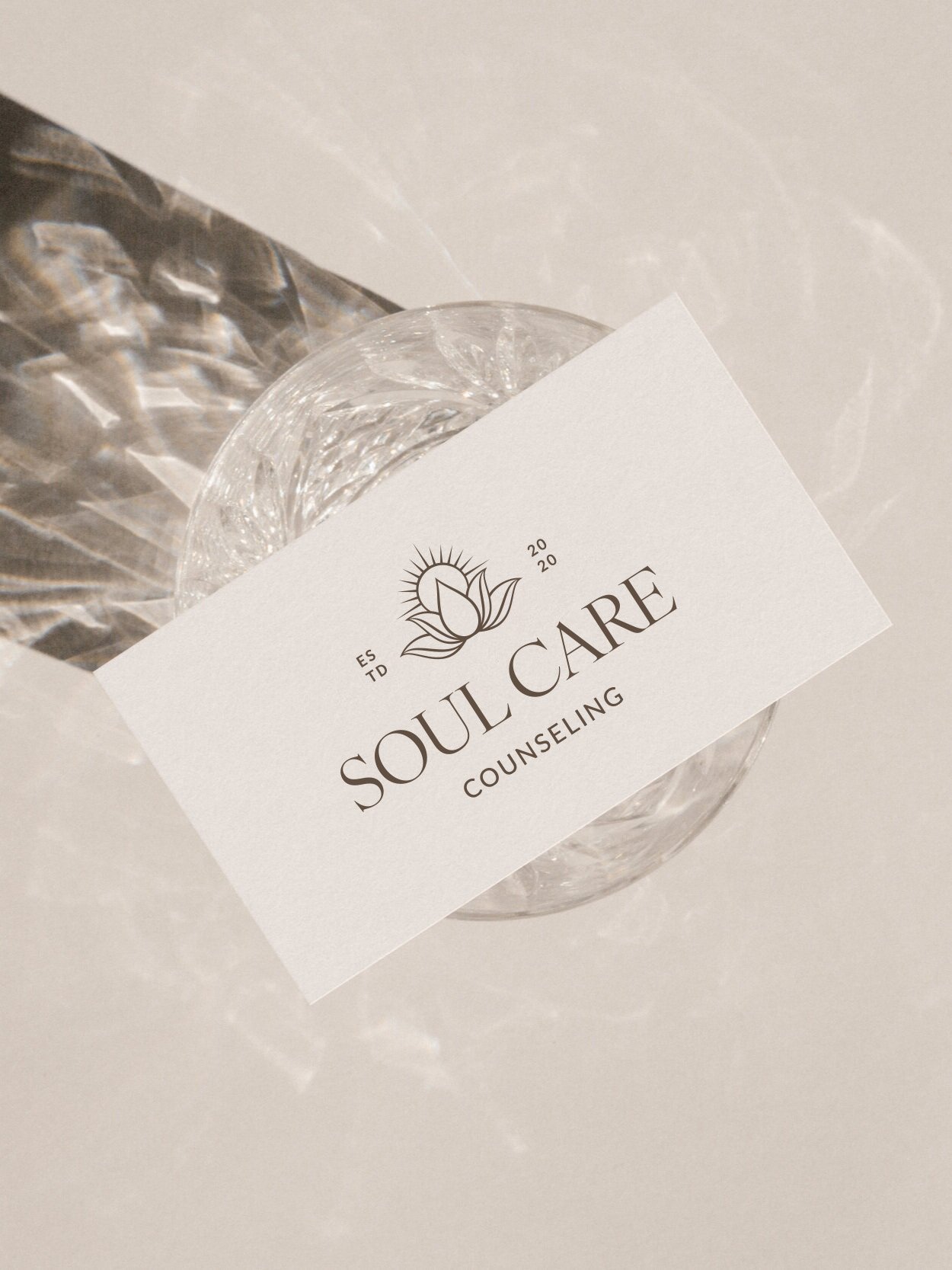 A business card for Soul Care Counseling placed on a glass of water with a decorative silver surface nearby, on a white table.