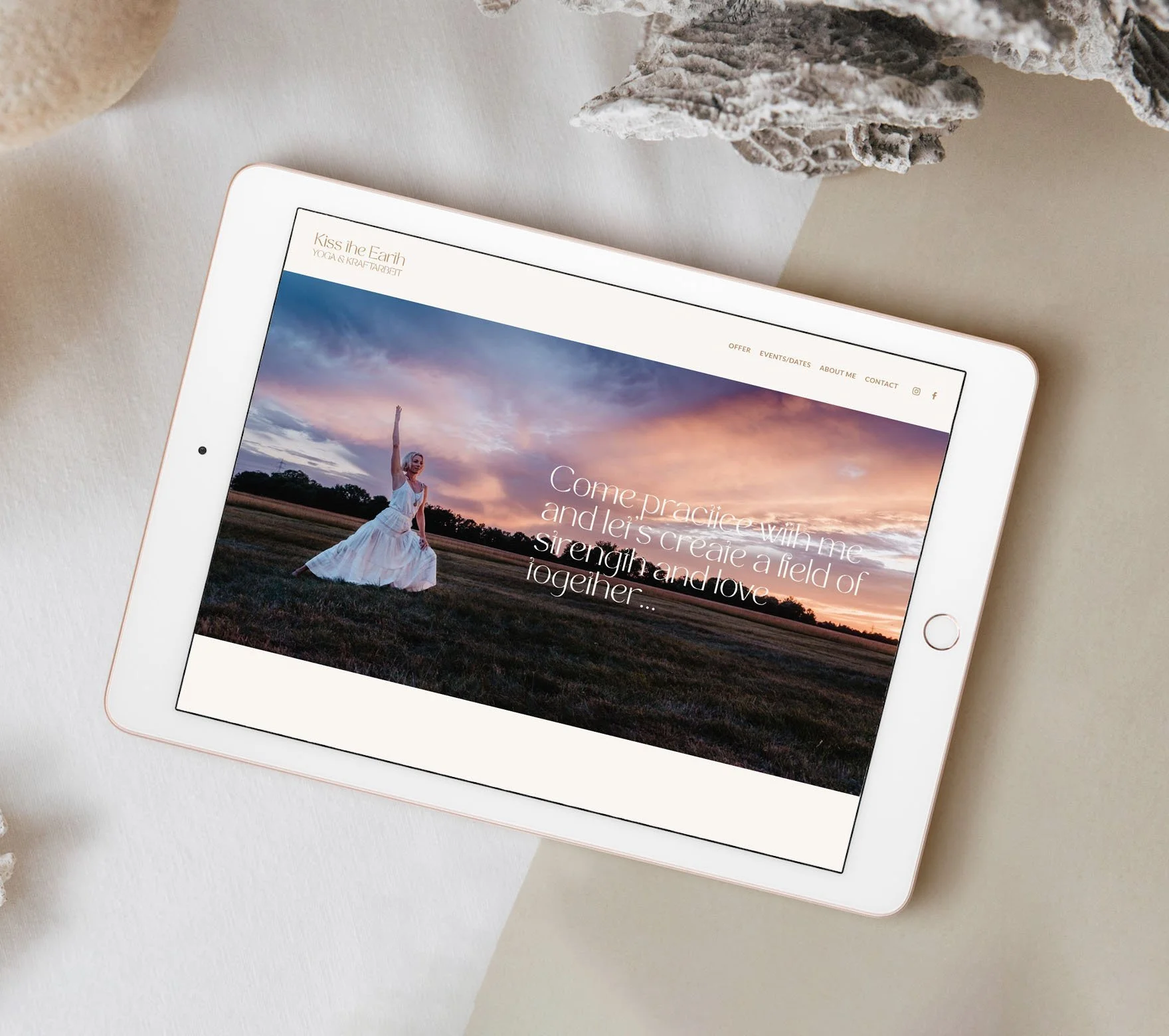 An iPad on a white surface displaying a website with a large background photo of a woman in a white dress standing in a field at sunset, with overlaid text inviting to practice yoga and create a field of strength and love together.