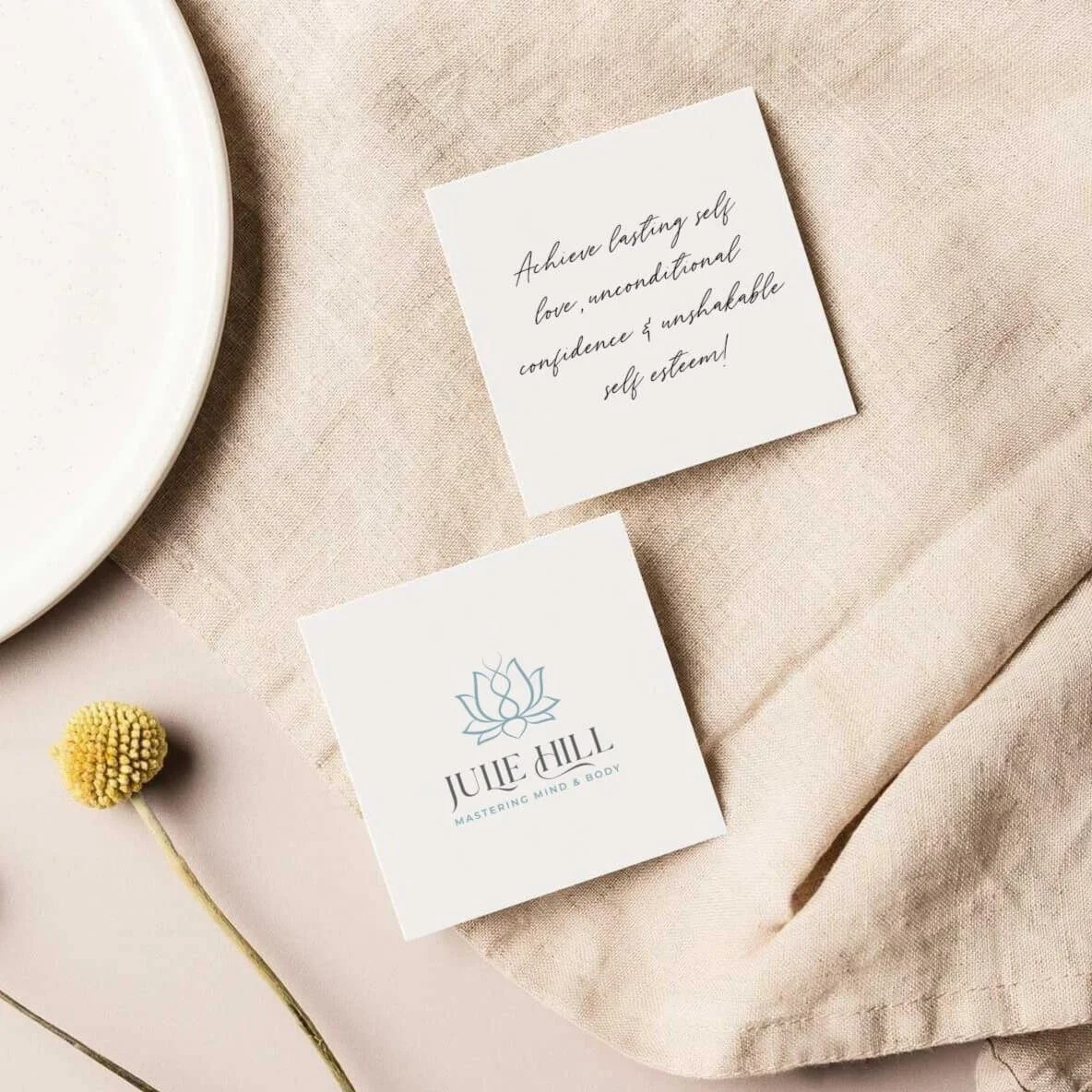 A white square card with a lotus flower logo and on a beige fabric background, along with a handwritten note about self-love and confidence, and a small yellow flower.