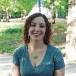 A woman with brown curly hair and teal blouse against outdoors background