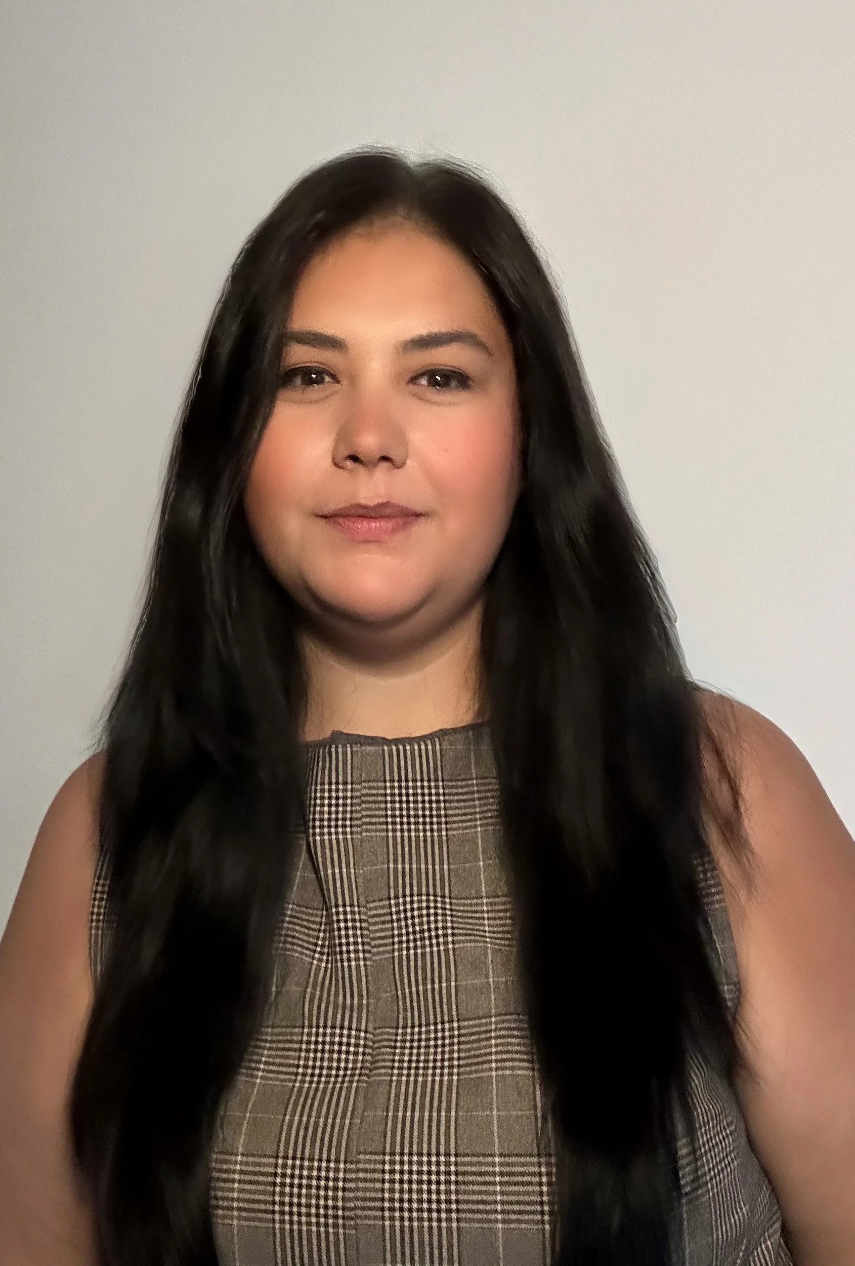 A portrait of a woman with long black hair, wearing a brown plaid sleeveless top, standing against a plain white wall.