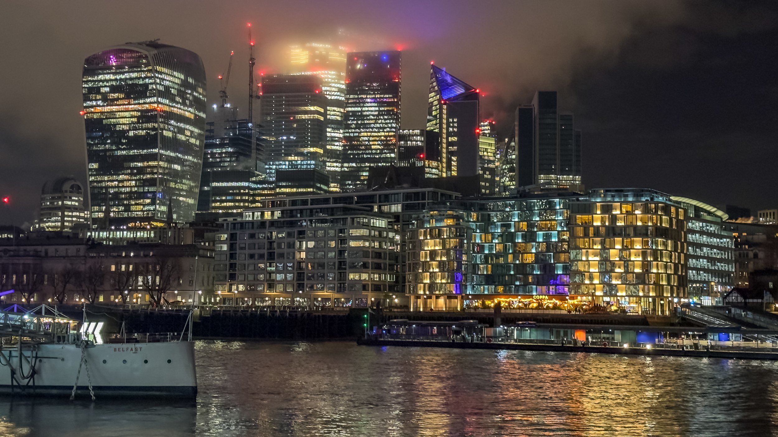 Night cityscape featuring modern illuminated buildings along the waterfront with reflections on the water, fog partially covering the taller skyscrapers.