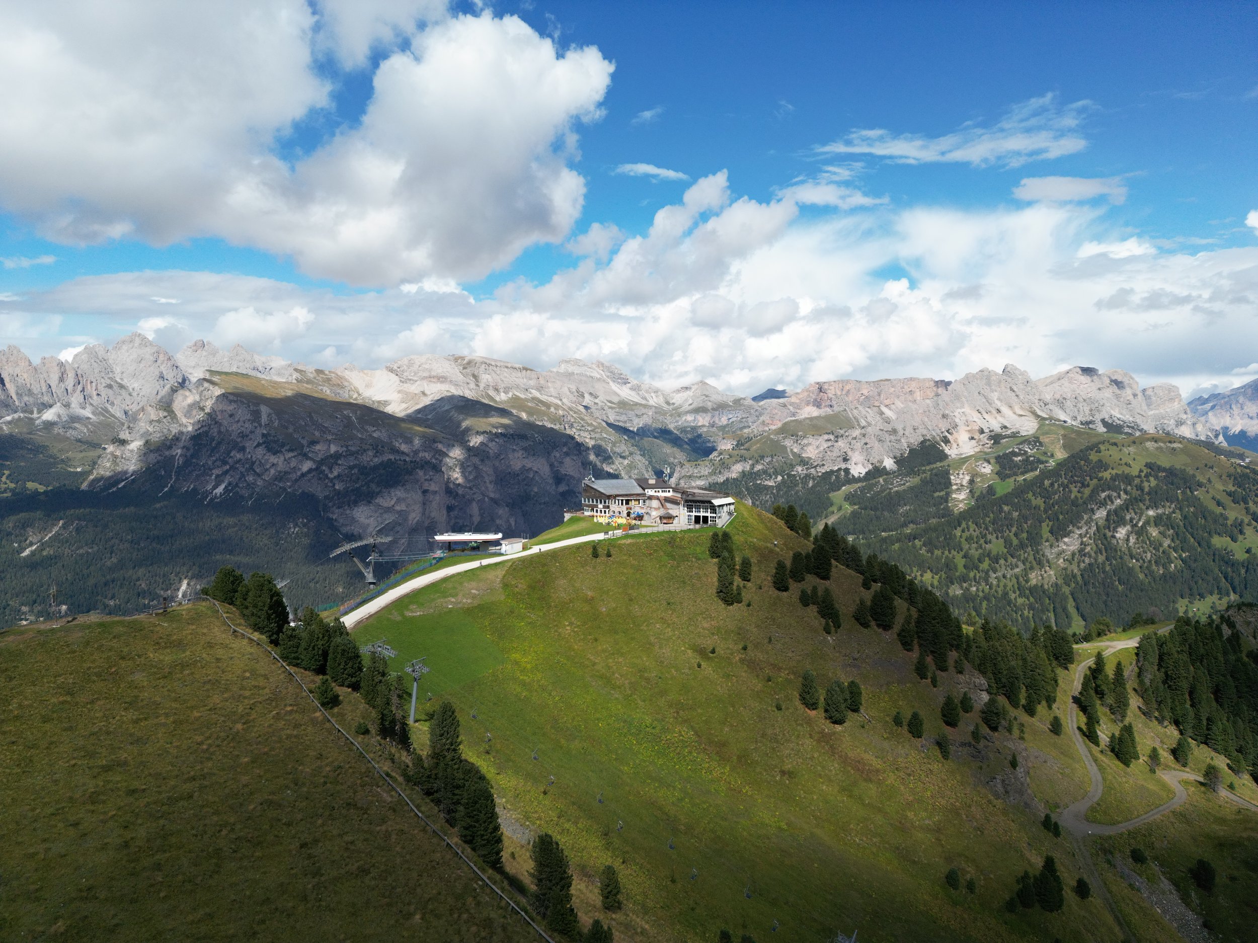 Green hillside with a modern building and ski lift chairs, set against mountains and a cloudy sky.