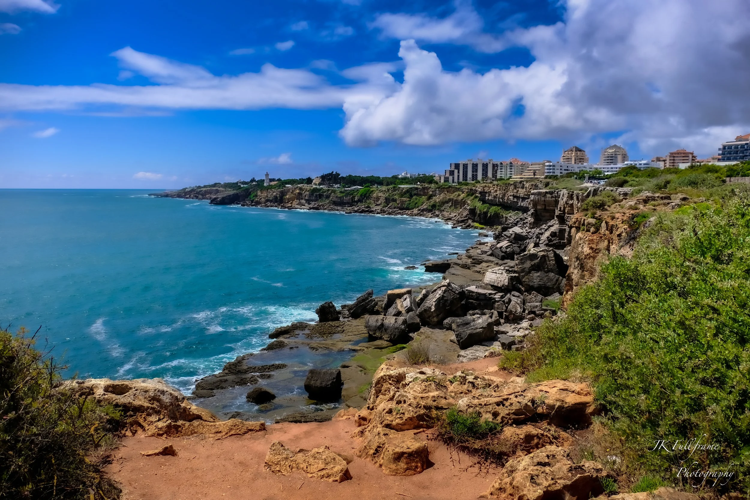 Coastal view of rocky cliffs overlooking the ocean, with a blue sky and clouds above, in a cityscape setting.