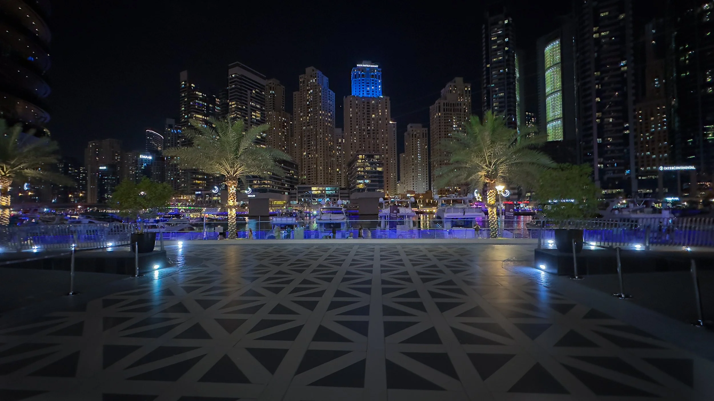 Nighttime view of a marina with yachts docked, surrounded by illuminated palm trees and tall city skyscrapers, some lit in blue and green lights.