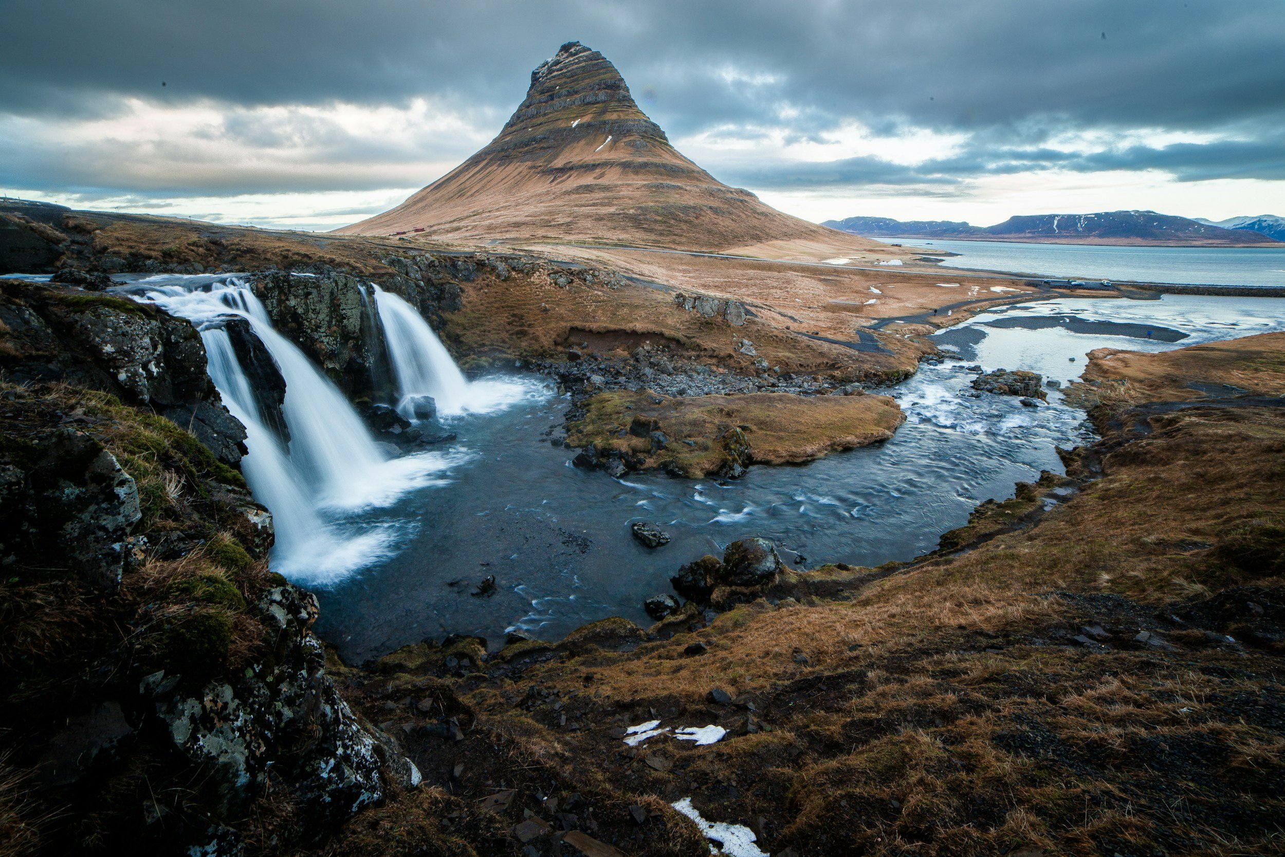 Scenic landscape of Kirkjufell mountain with cascading waterfalls and rushing river in Iceland during overcast weather.