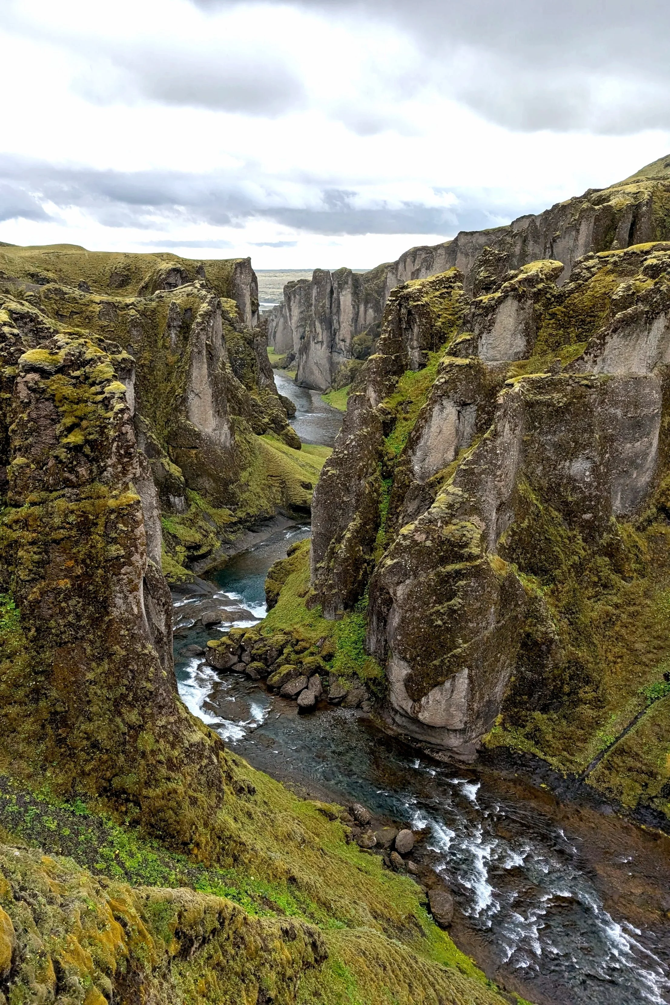 A river flowing through a deep canyon with moss-covered rocky walls, under a cloudy sky.