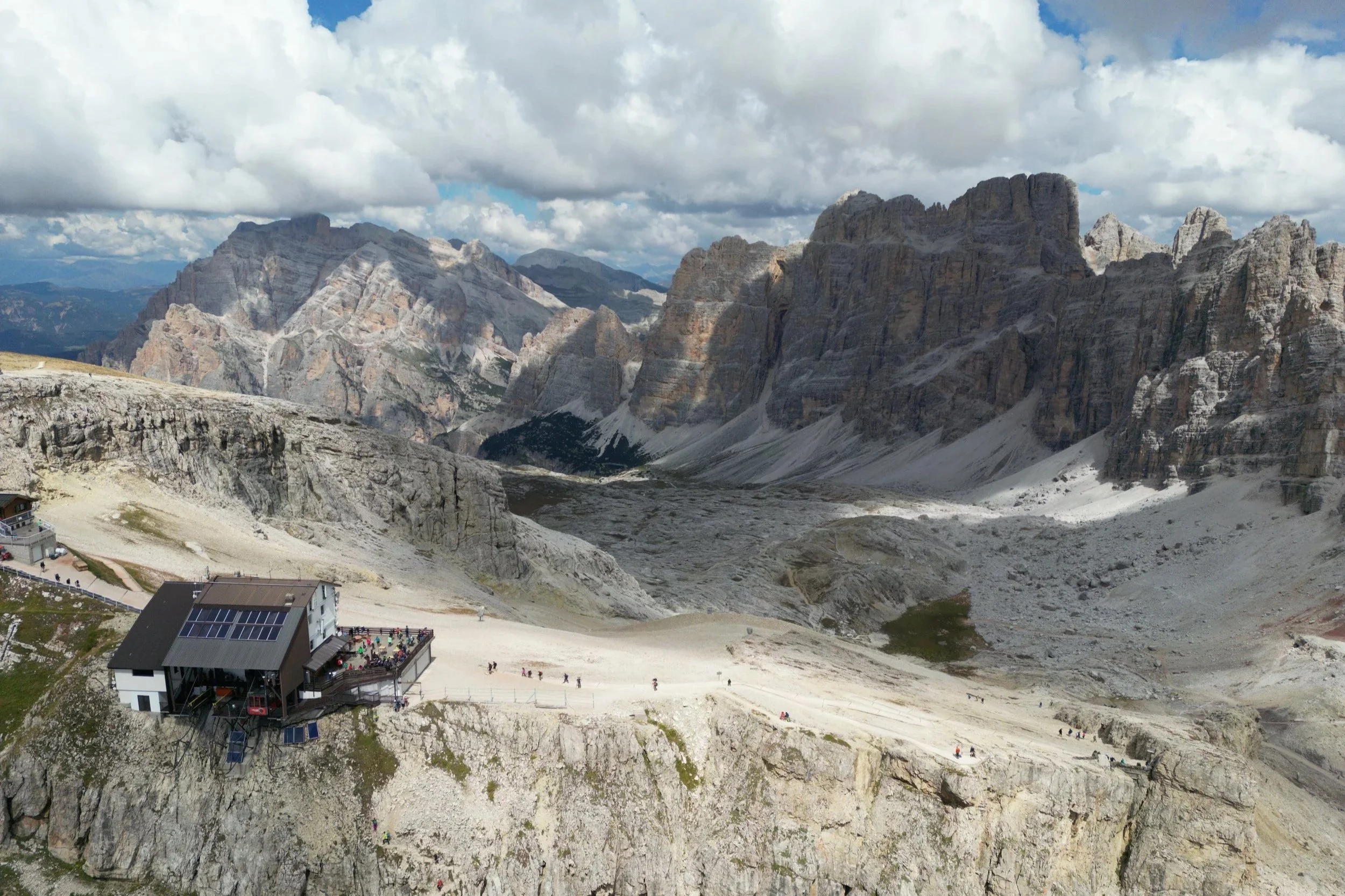Mountain landscape with rocky cliffs and peaks under partly cloudy sky, featuring a building and tourists on the mountain ledge in the foreground.
