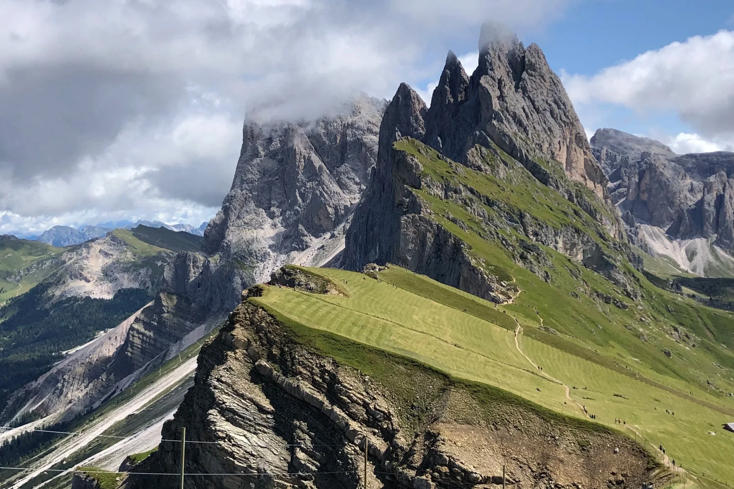 Scenic view of mountain range with sharp peaks partially covered by clouds, green grassy slopes, and a winding trail.