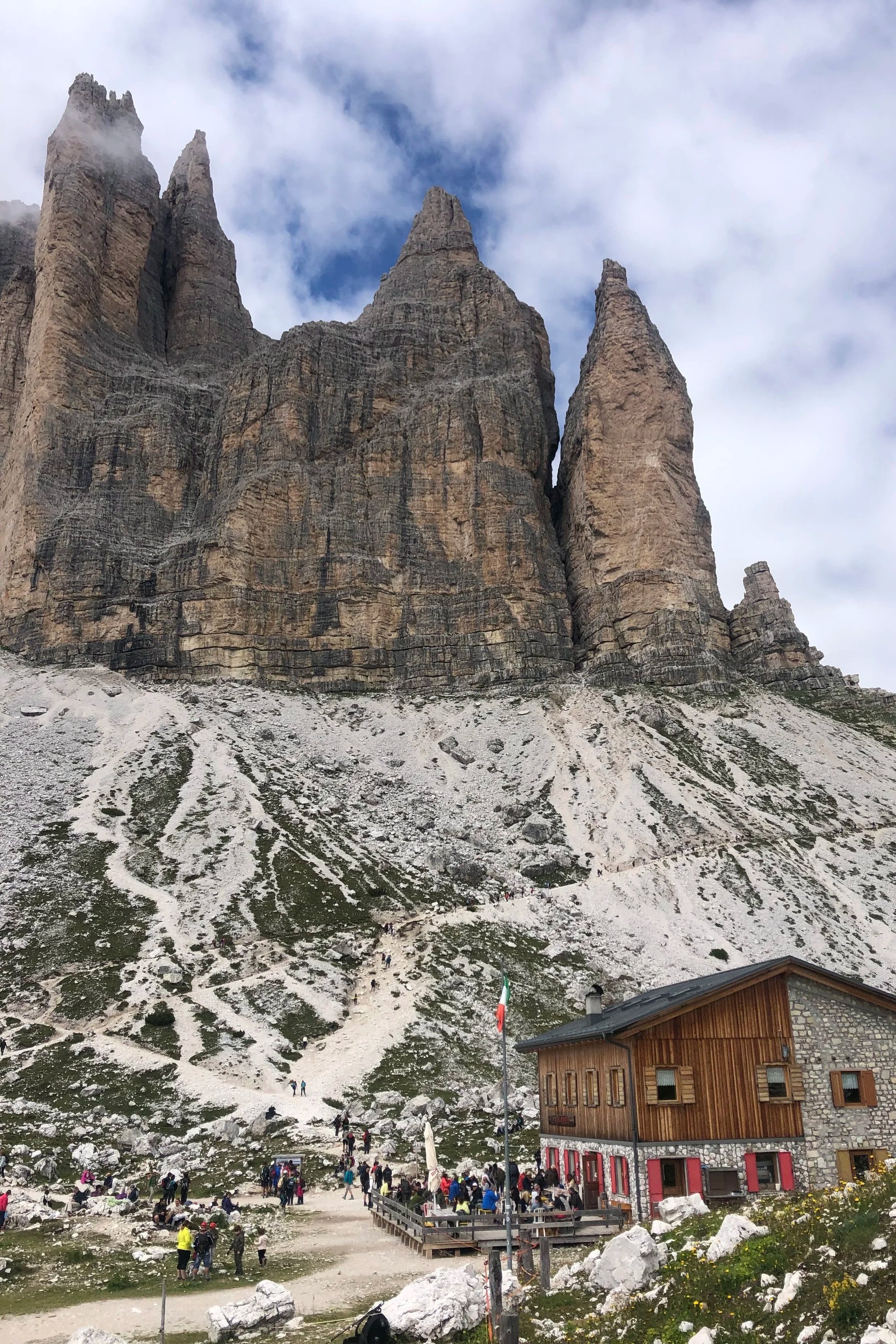 A mountainous landscape with towering rocky cliffs and a rocky slope below. There is a wooden building with red accents at the base, and many people are gathered around, some walking up the trail and others near the building.