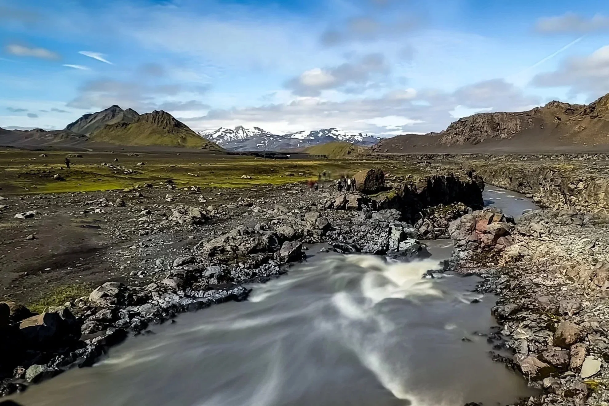 A rocky river flowing through a valley with mountains in the background, some with snow caps, under a partly cloudy sky.