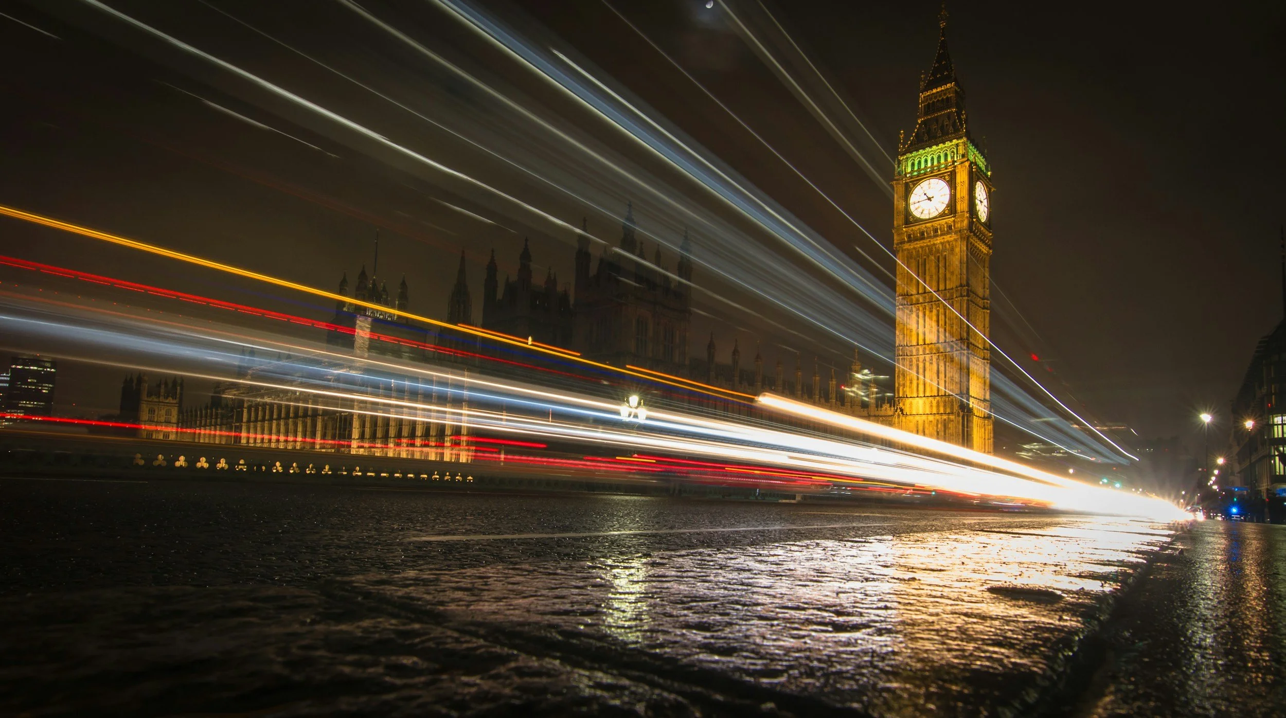 A nighttime scene of Big Ben clock tower in London with streaks of light from passing vehicles across the foreground, and the Palace of Westminster in the background.