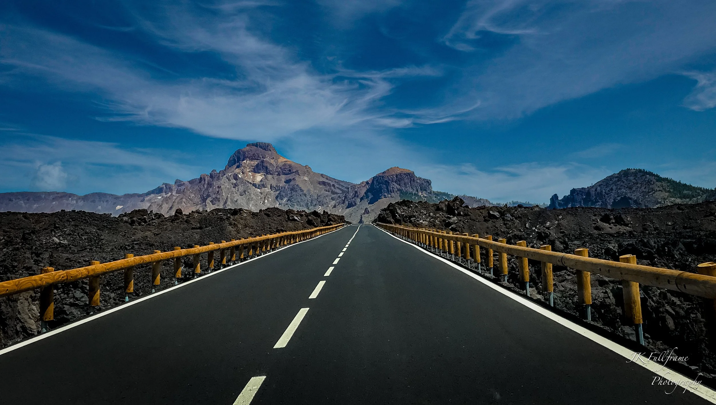 A straight paved road with yellow guardrails on both sides, leading towards distant mountains under a partly cloudy blue sky.