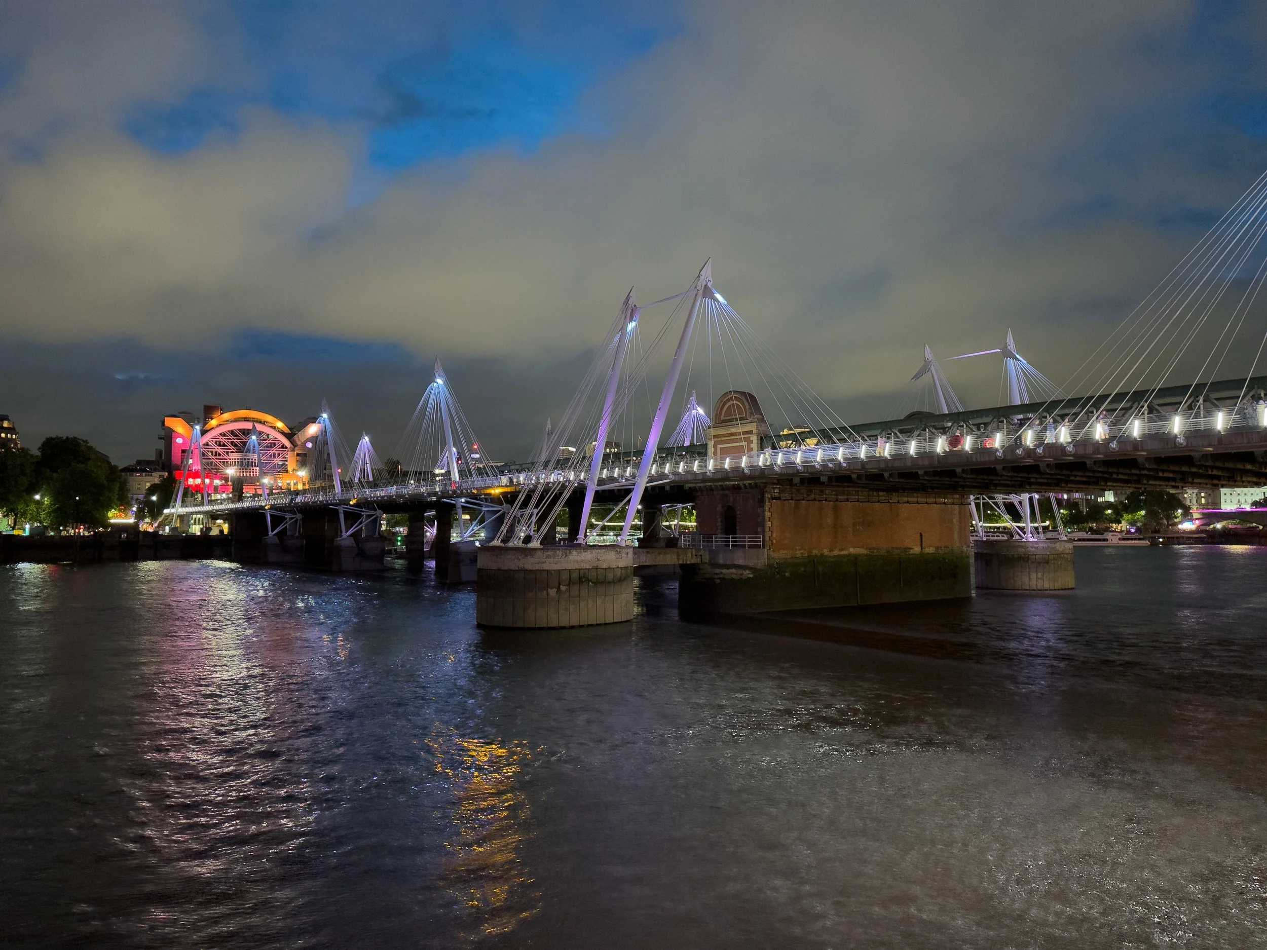 Night view of a modern bridge over a river, illuminated with purple and white lights, with a city skyline and a lit Ferris wheel in the background.