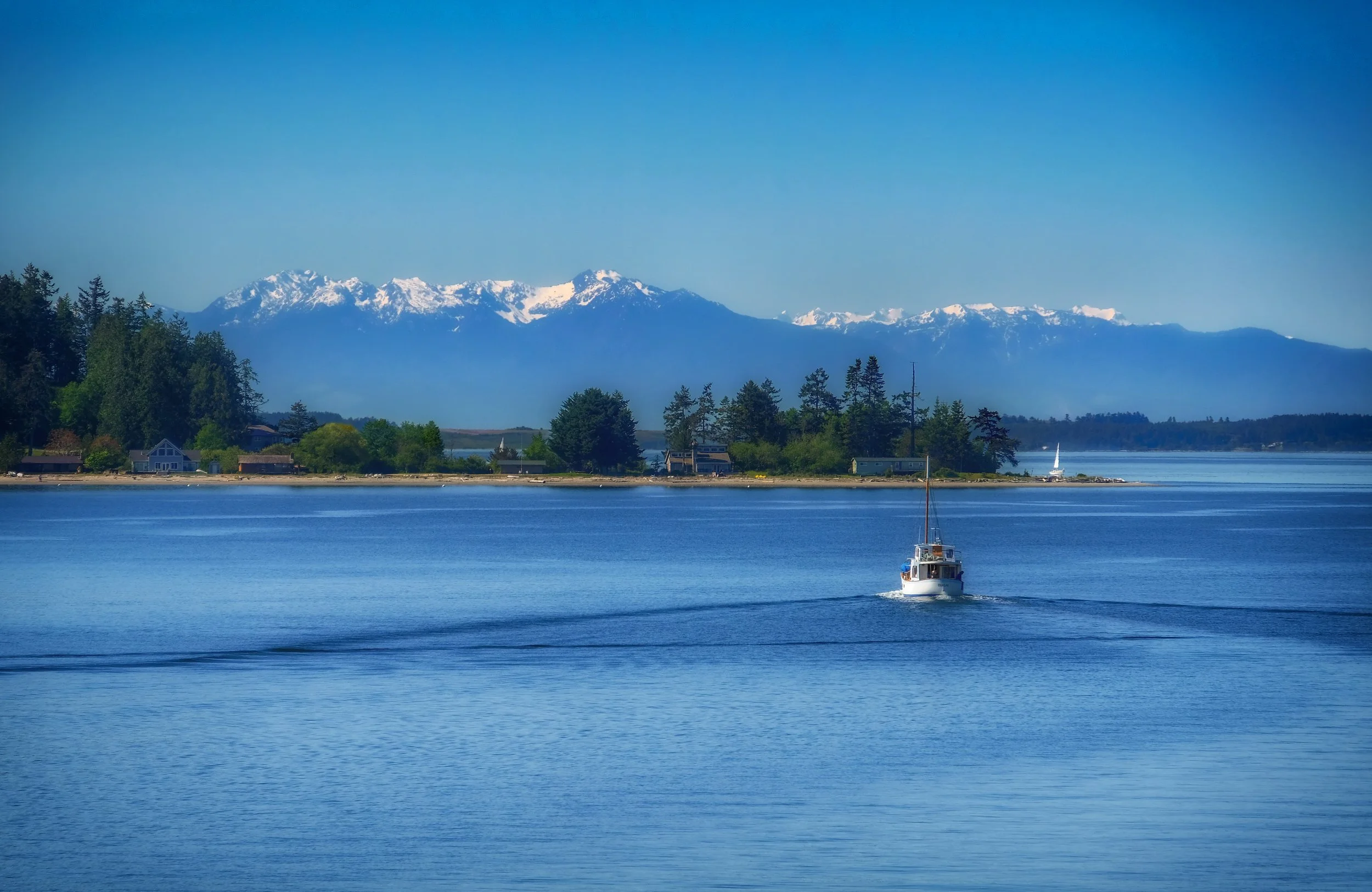 A boat sailing on a calm body of water with a small shoreline and houses in the background, snow-capped mountains under a clear blue sky.