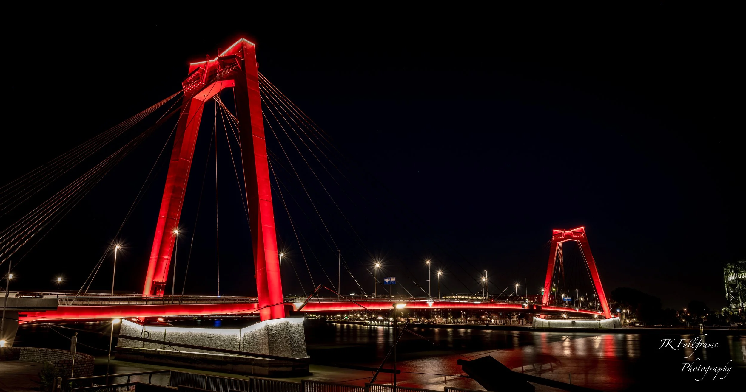 Nighttime view of a modern cable-stayed bridge illuminated in red, spanning over a calm body of water, reflected on the surface.