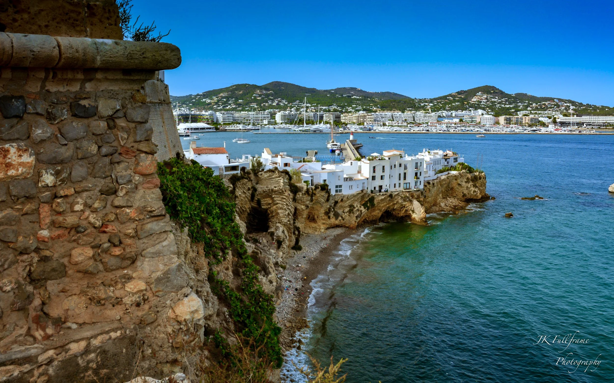 A coastal scene with white buildings on rocky cliffs overlooking the sea, with sailboats in the harbor and green mountains in the background.