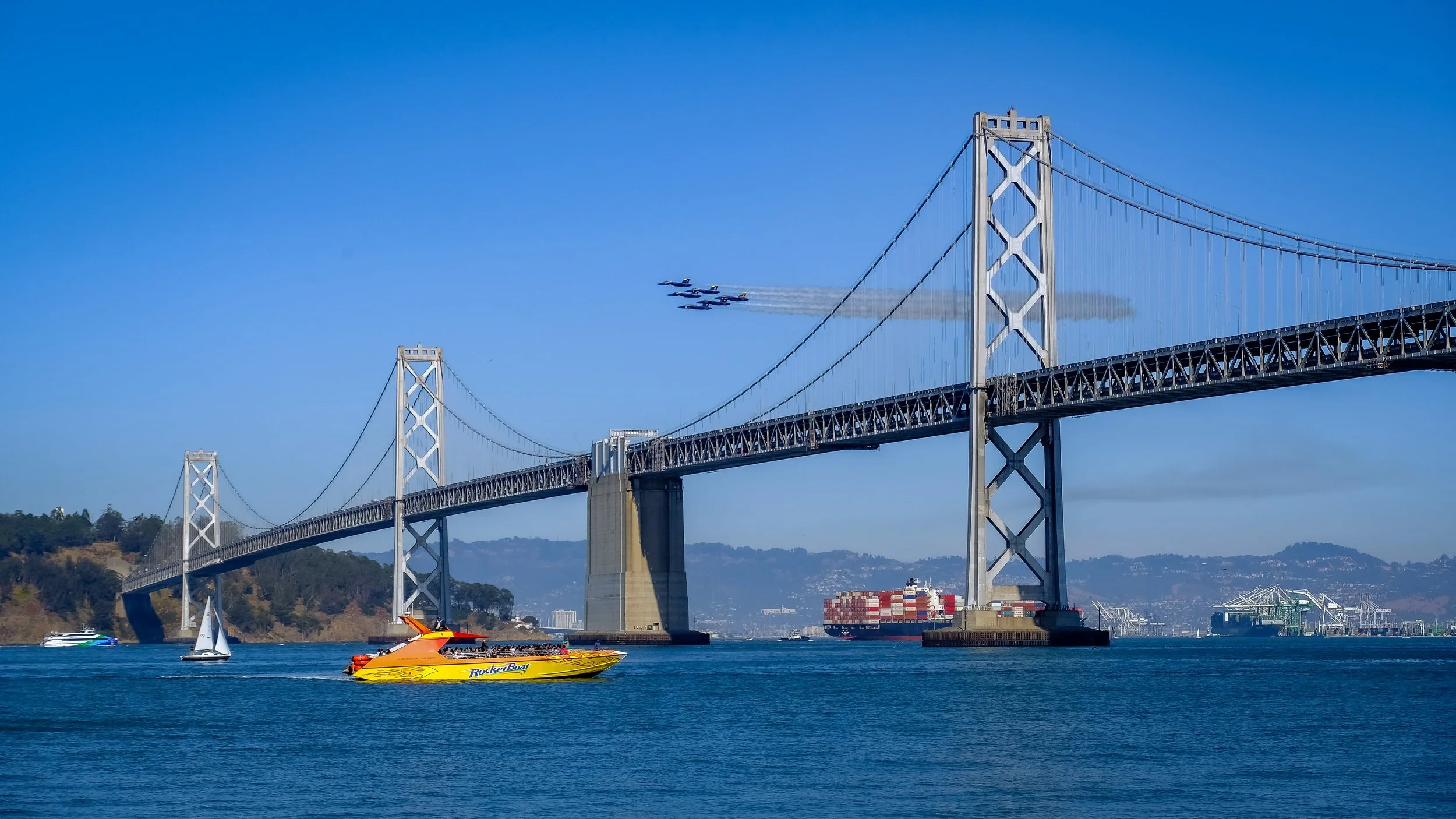 A large suspension bridge over a body of water with boats, including a yellow speedboat and sailboats, and planes flying in formation in the sky.