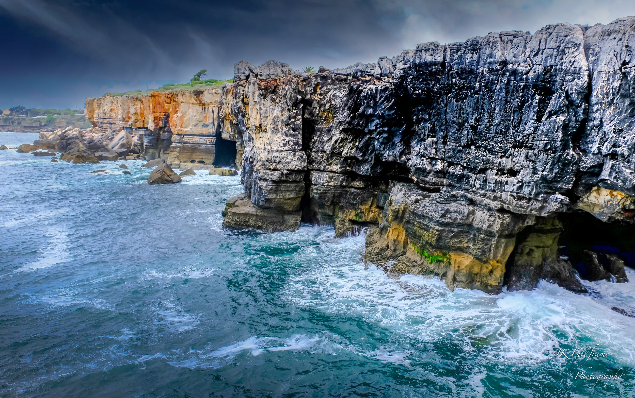 Rocky ocean coastline with cliffs, waves crashing against rocks, overcast skies.