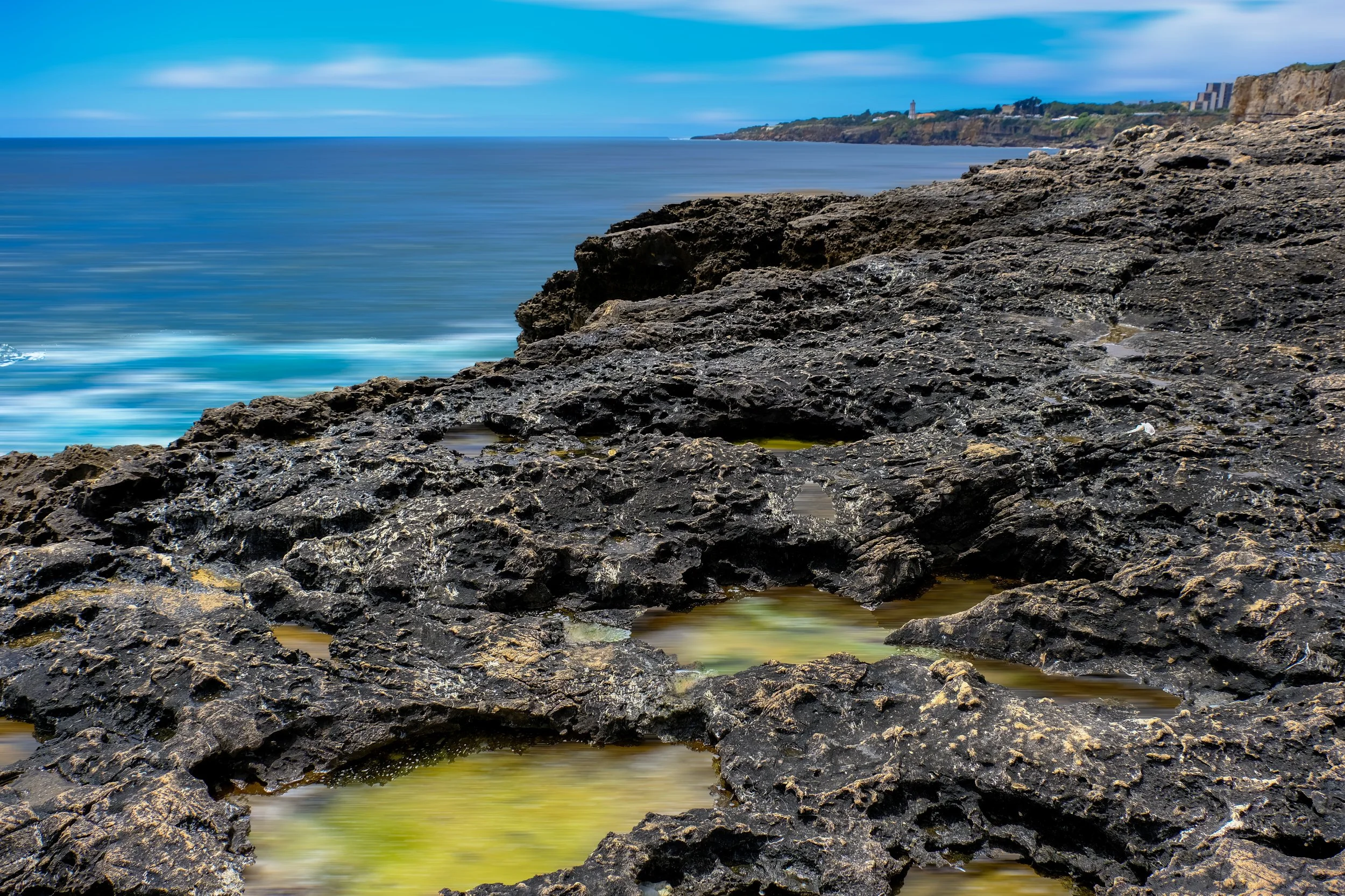 Rocky shoreline with tide pools in the foreground, ocean waves in the middle, and a distant coastline with buildings and a lighthouse under a cloudy sky.