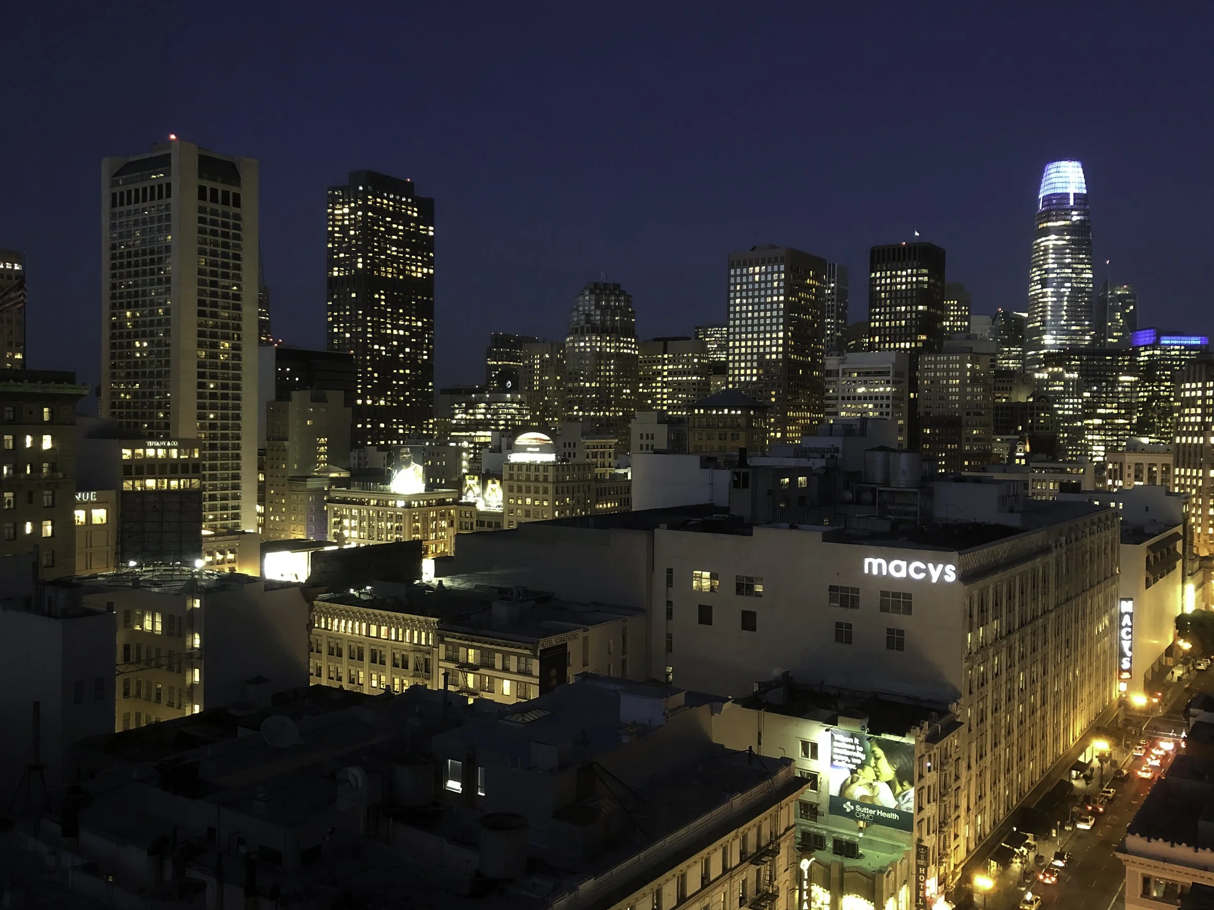 Nighttime cityscape of San Francisco with illuminated skyscrapers, including Salesforce Tower, and Macy's building in the foreground.