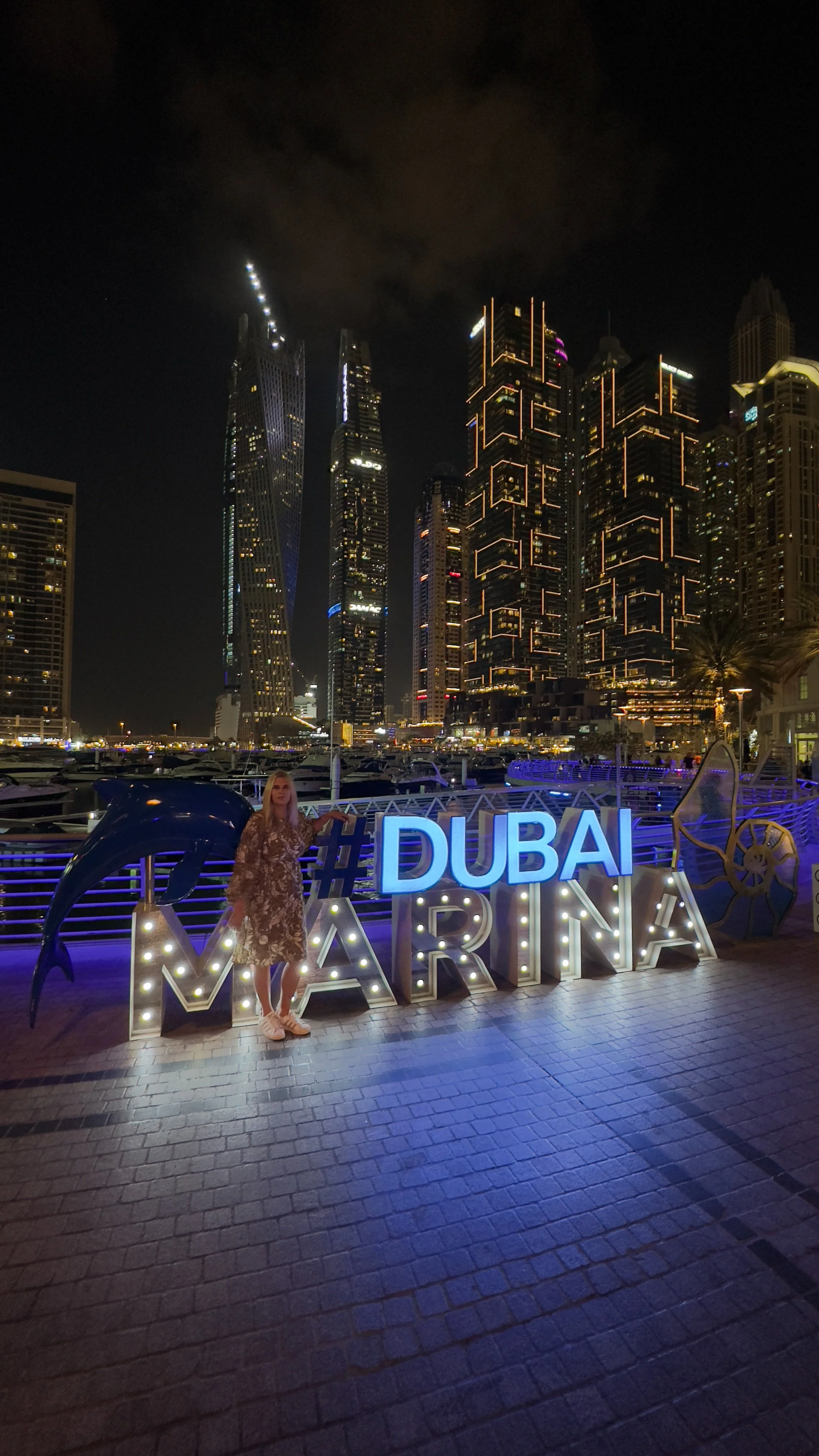 Nighttime view of Dubai Marina skyline with tall illuminated skyscrapers and a woman standing beside a sign that reads '#DUBAI MARINA' with lights, near the water with boats in the background.
