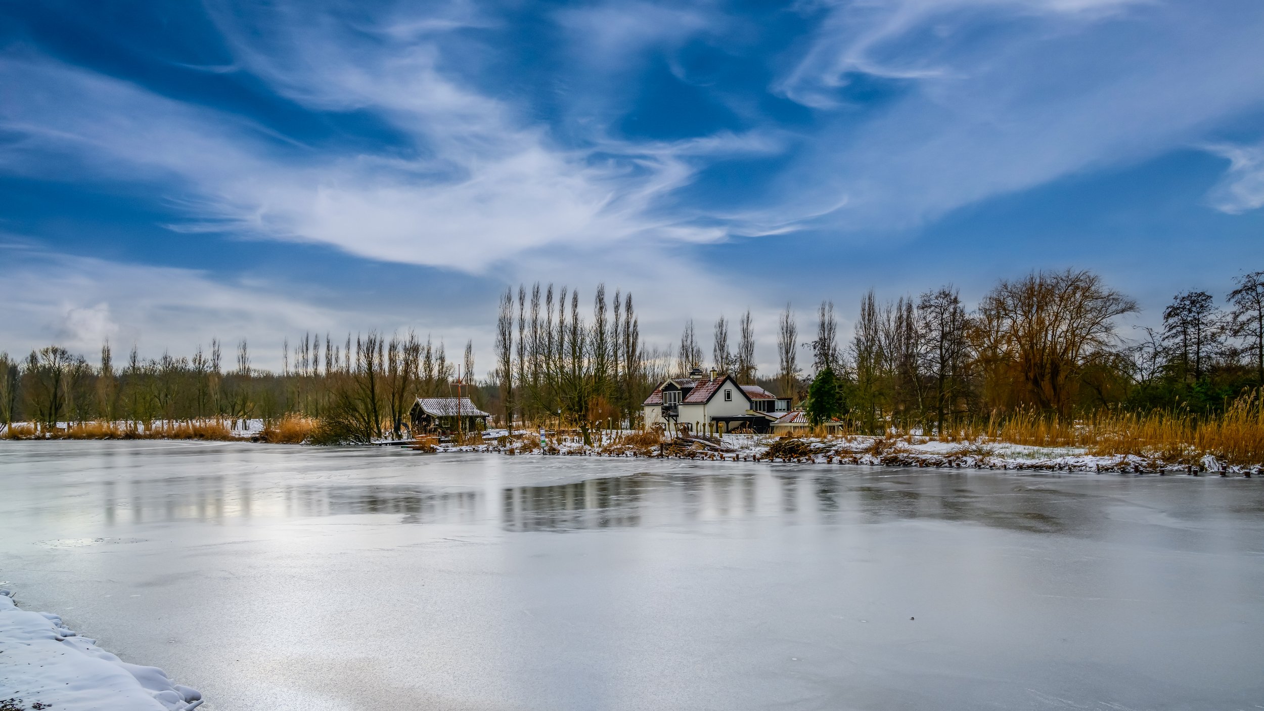 A frozen lake in the Netherlands Midden Delfland during winter with snow on the shore and trees in the background, under a partly cloudy blue sky.