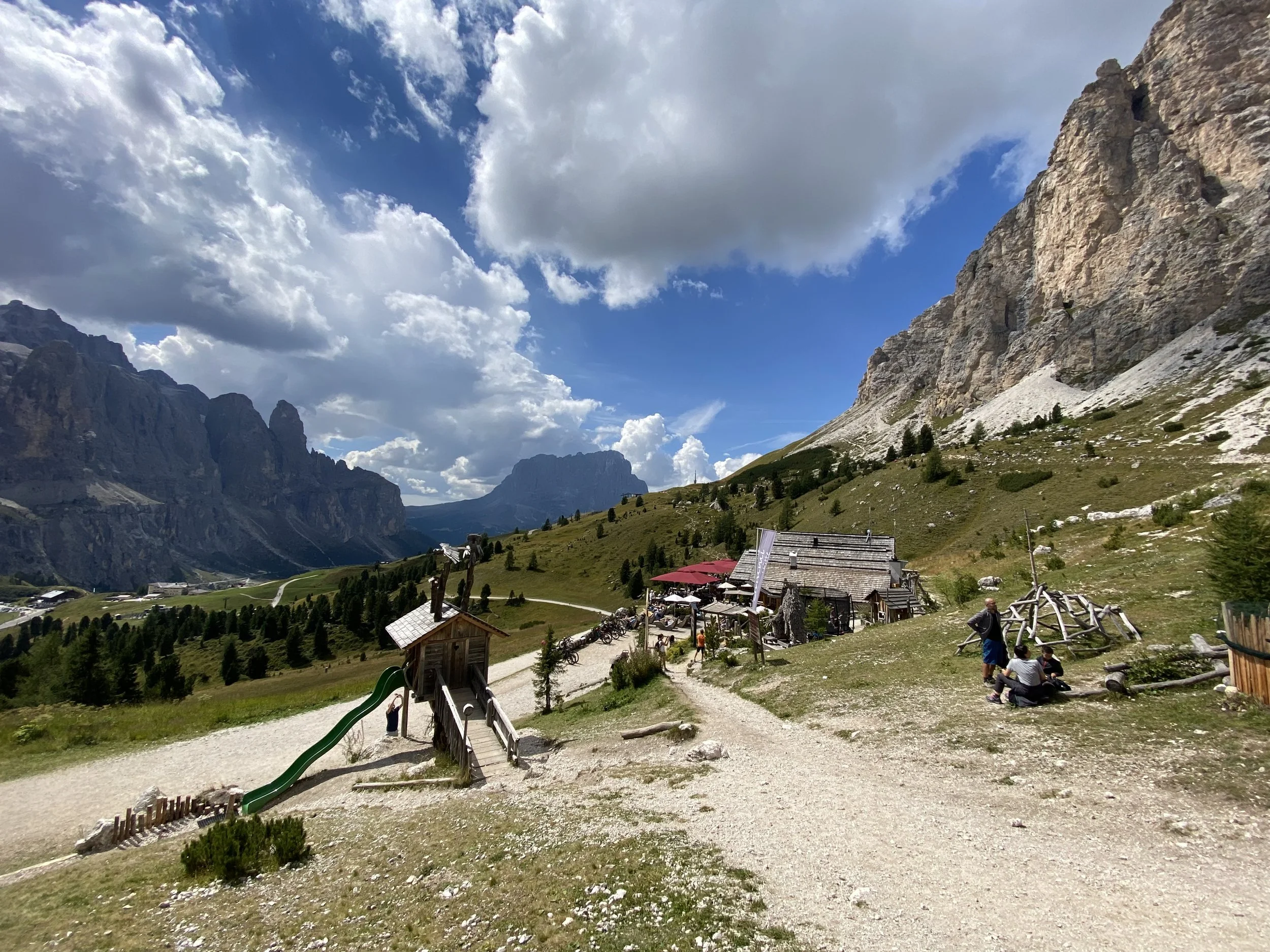 Mountain landscape with a small rustic building and people sitting outside. Dirt paths wind through green grassy areas with tall mountains and dramatic clouds in the background.