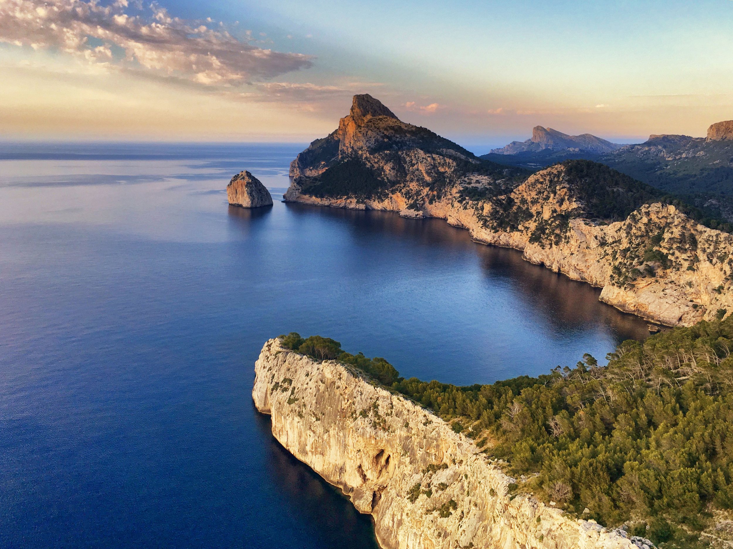 Aerial view of a coastal landscape with a large rocky island in the sea, surrounded by cliffs and green trees, under a partly cloudy sky at sunset.