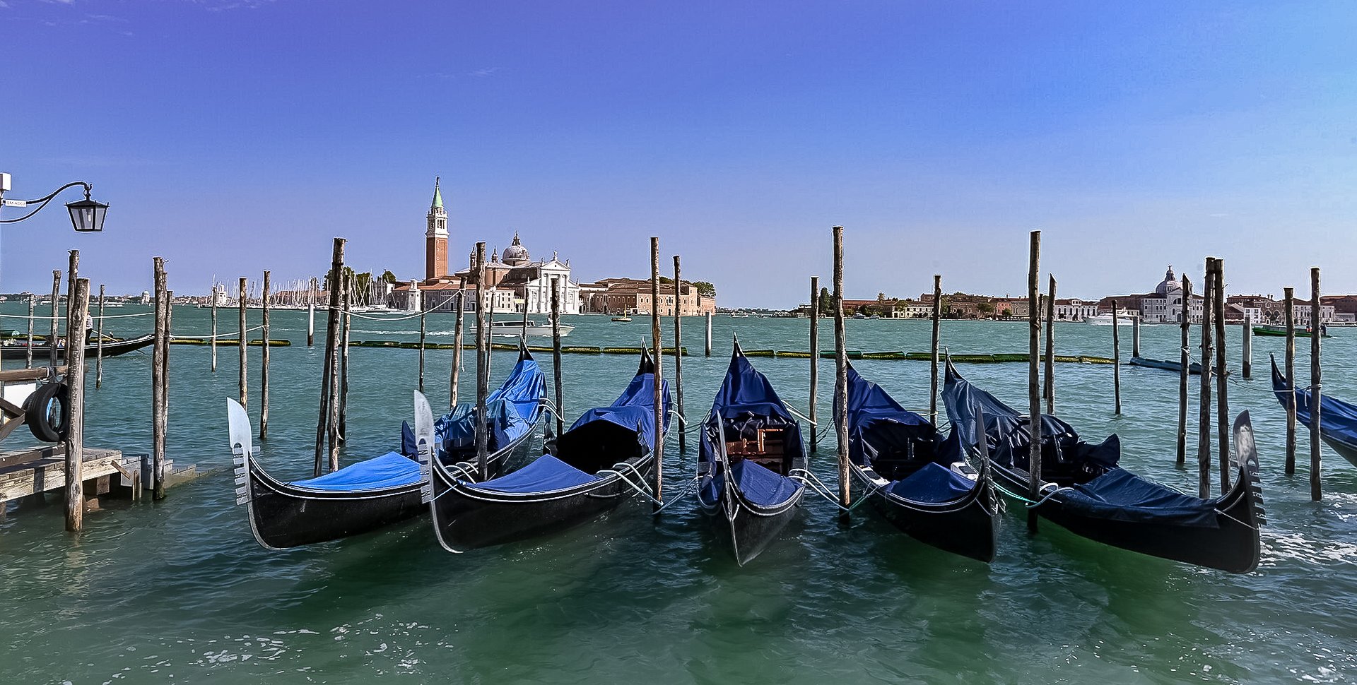 Gondolas docked along a pier in Venice with historic buildings and church towers in the background under a clear blue sky.