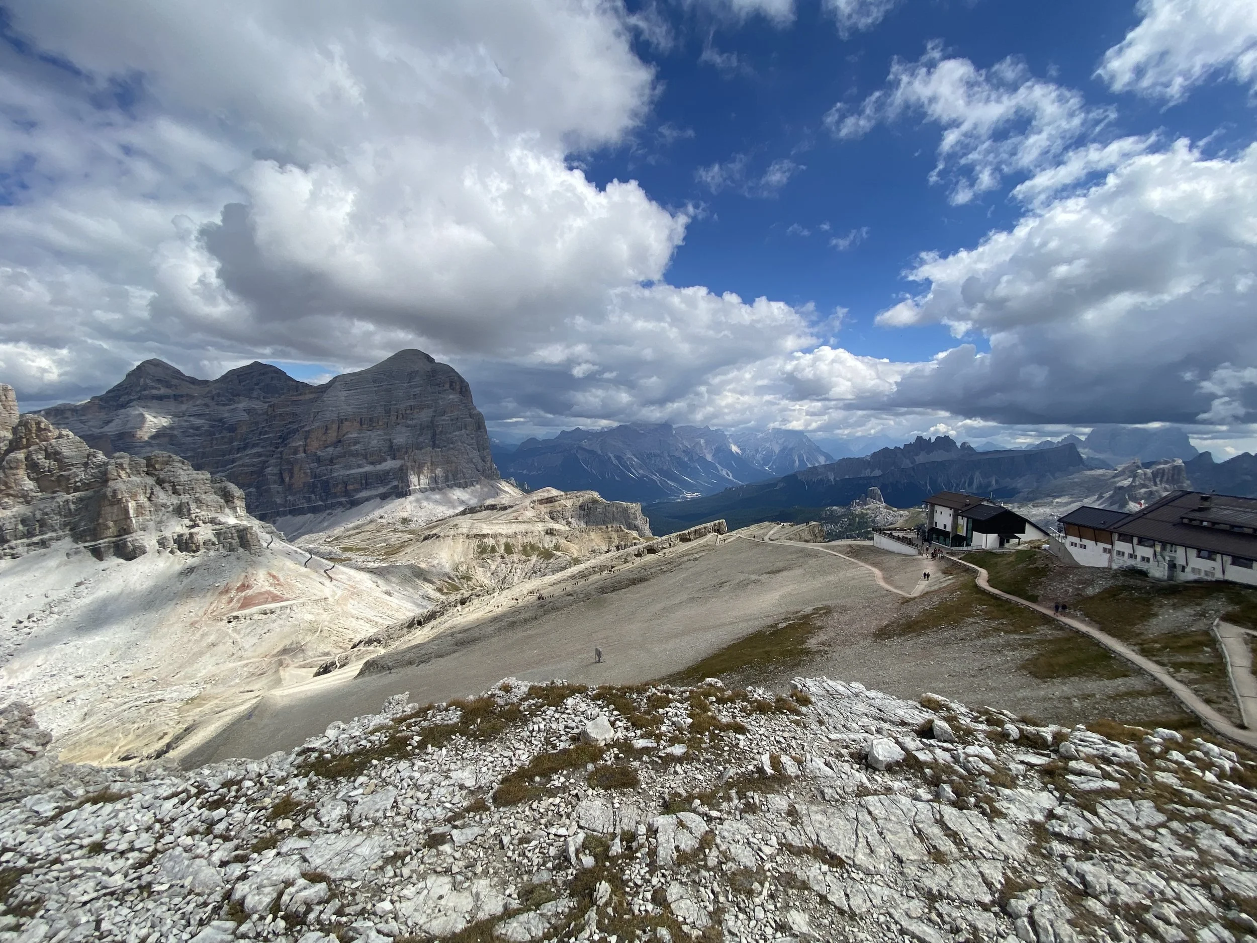 Mountain landscape with rocky terrain, mountainous peaks, a partly cloudy sky, and a few buildings along a winding pathway.
