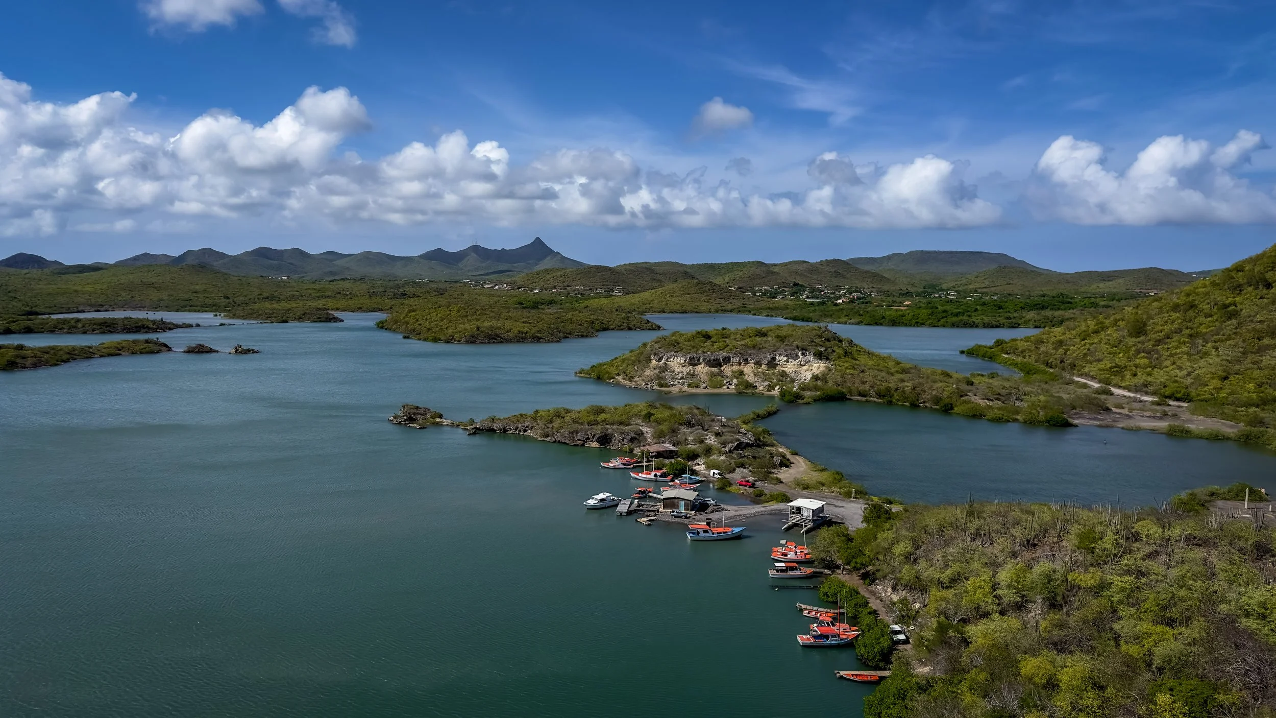 A scenic landscape with a river or bay surrounded by green hills and mountains under a partly cloudy sky.