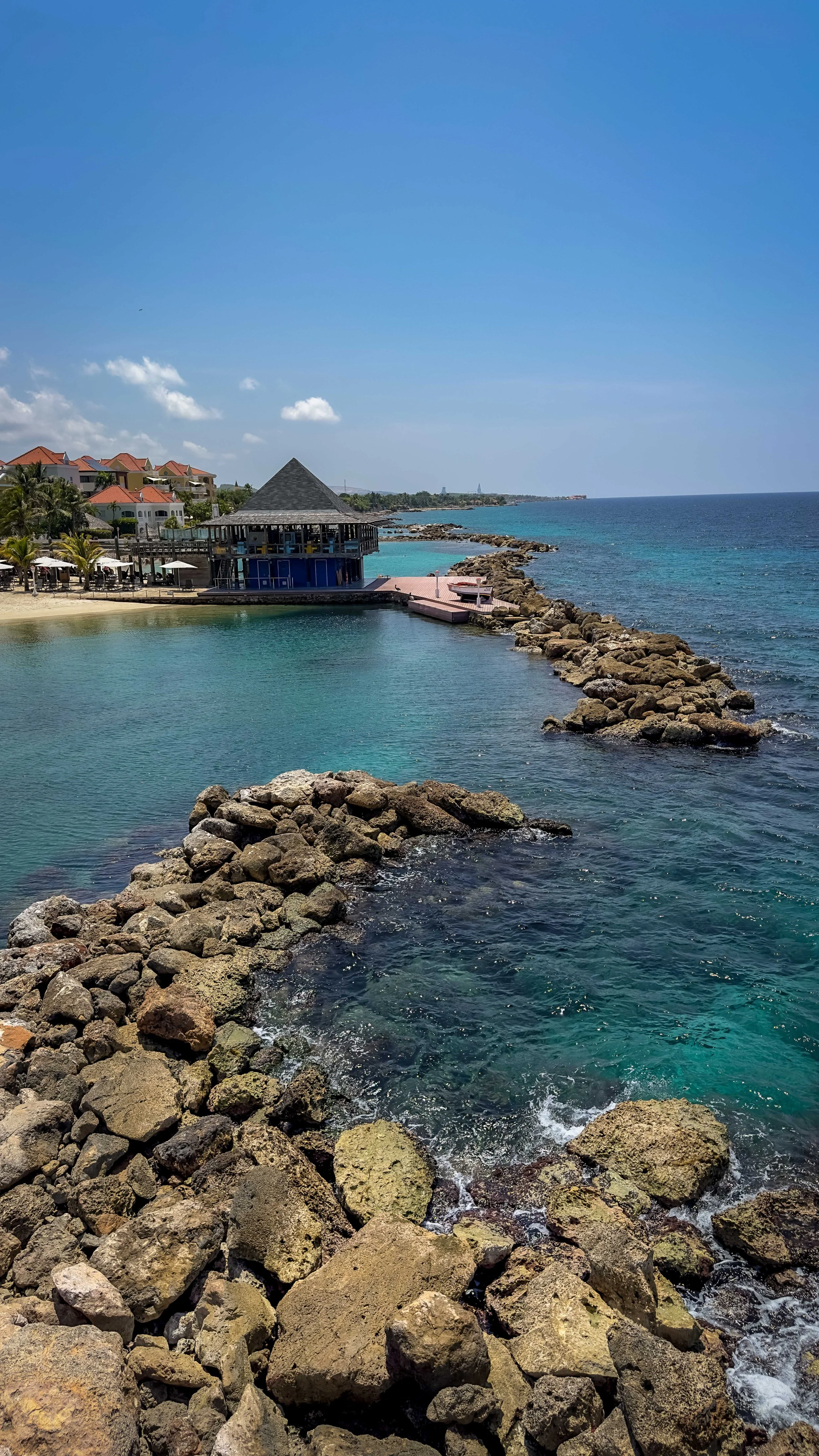 View of a rocky pier extending into the ocean near a pool area with beachside resort houses and a blue sky.