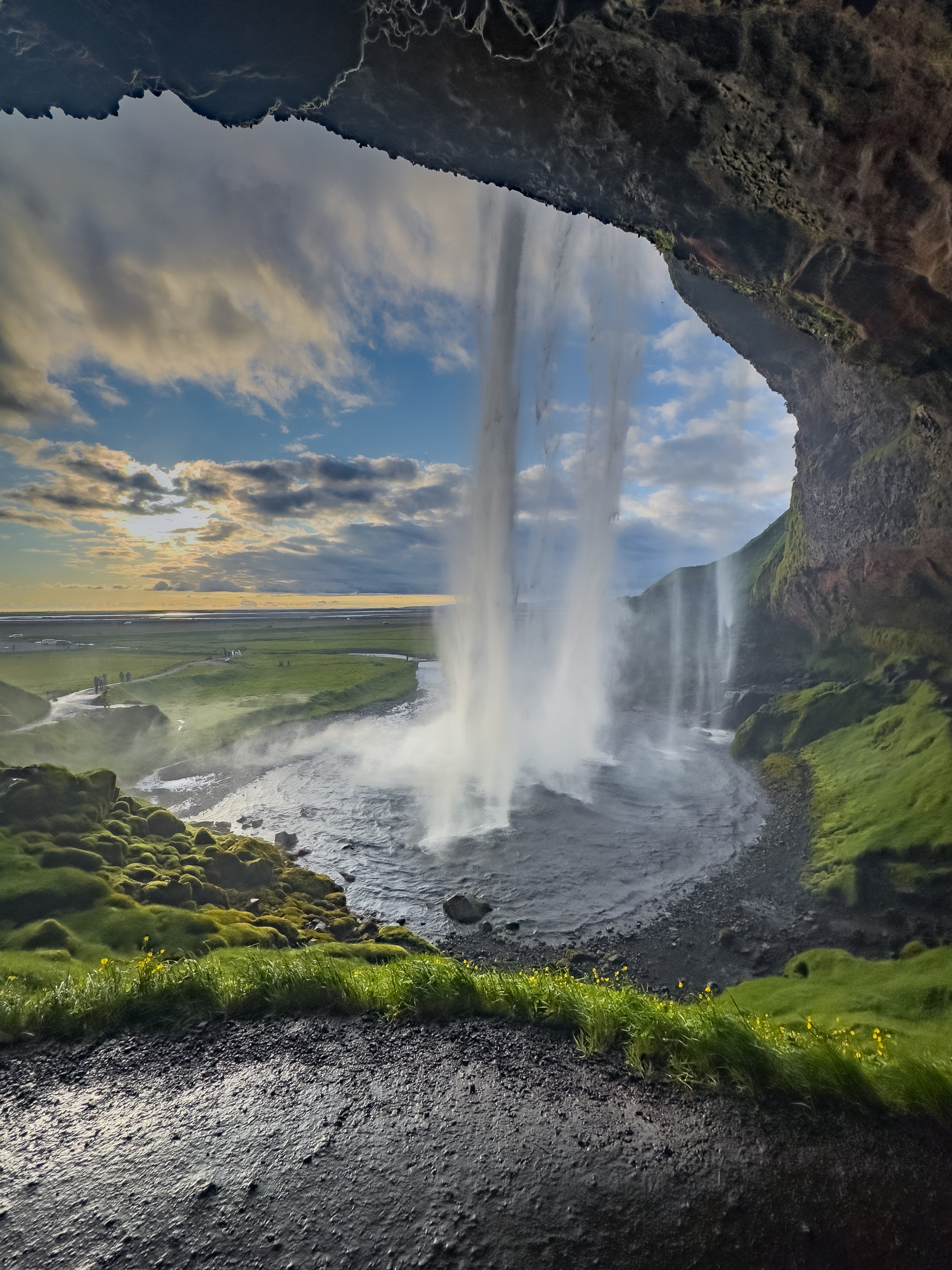 Stunning Seljalandsfoss waterfall in Iceland, a powerful curtain of water dropping into a pool, with lush green cliffs and people walking on the path that goes behind the falls