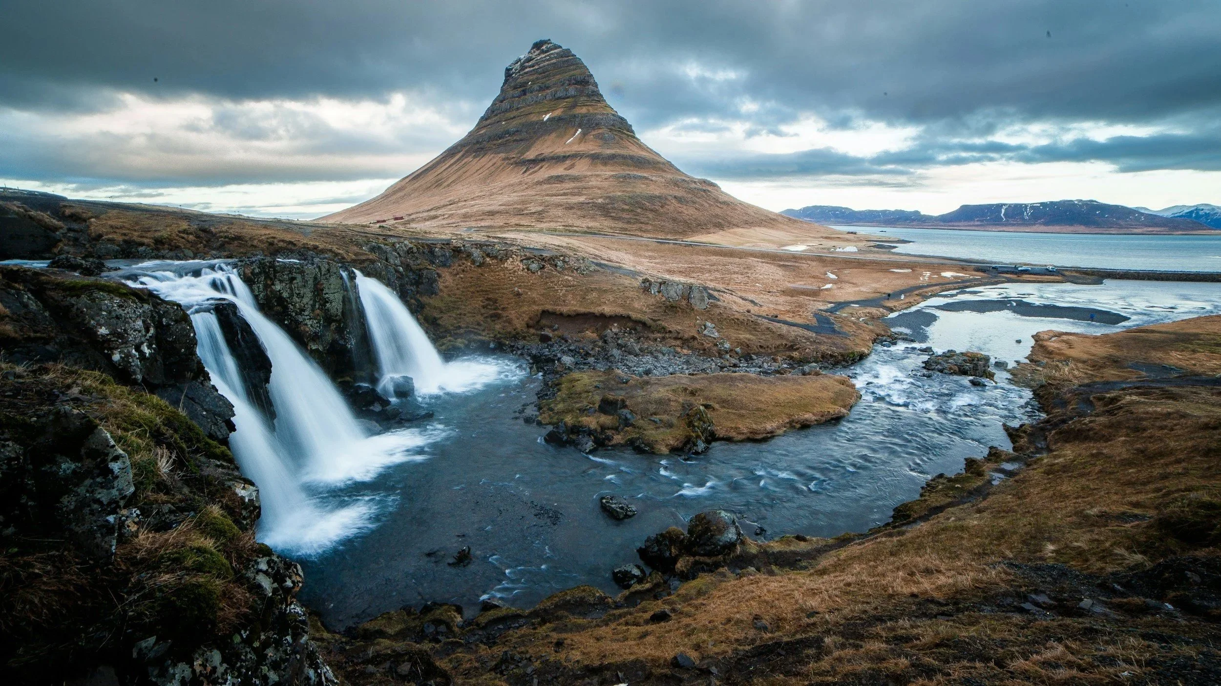 Scenic landscape featuring a waterfall with cascading water in the foreground, rugged terrain with brown and green hues, a prominent conical mountain in the background, overcast sky, and a body of water in the distance.