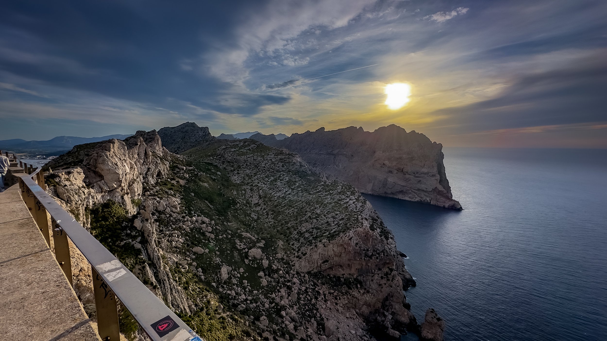Sunset over rocky cliffs and a calm ocean, with a viewing platform and railing in the foreground.