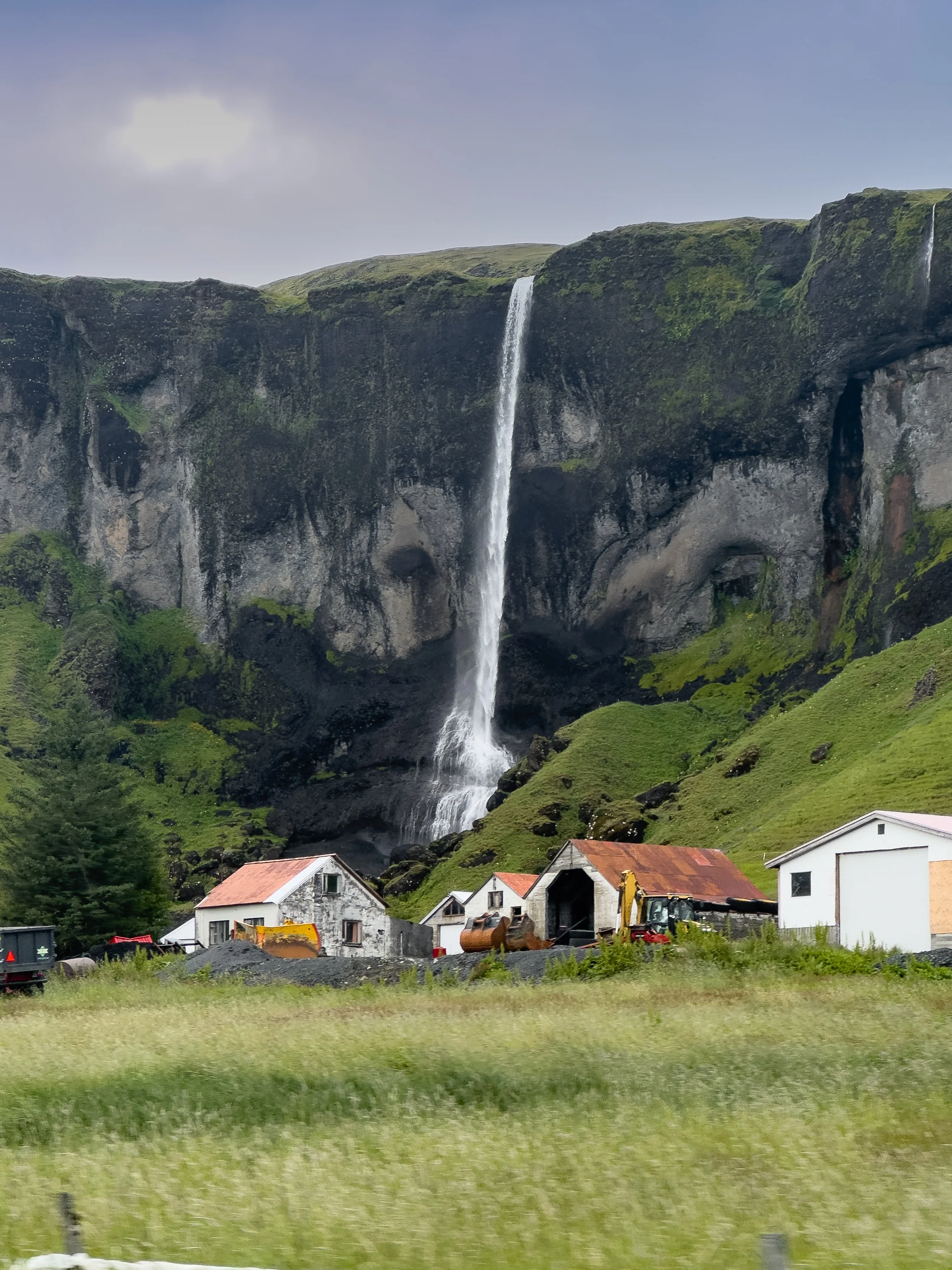 Ffarm buildings in front of a tall waterfall in along Iceland Ring Road cascading down a steep, moss-covered cliff in a rural area.